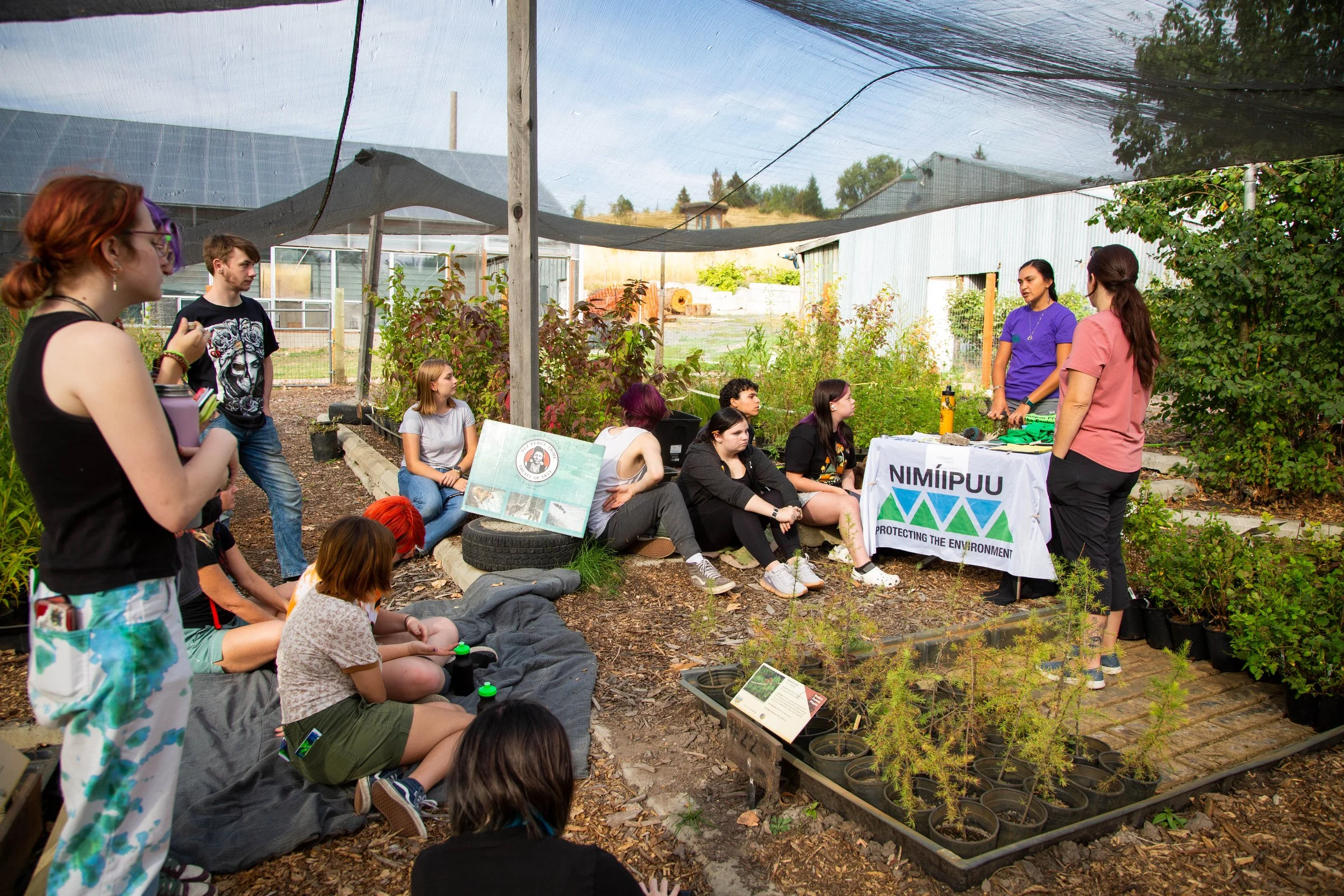 A group of young people attending an outdoor educational event at a garden or farm. Two women are standing and speaking at a table with a banner, while several others are seated or standing around, listening. The setting includes plants, trees, and a