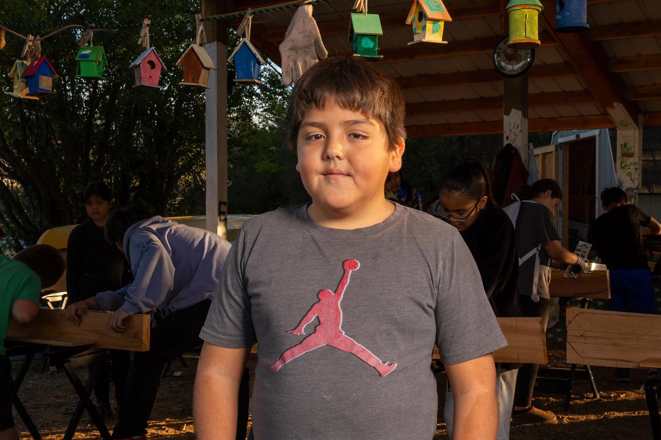 A young boy with brown hair wearing a gray T-shirt with a pink Jordan logo, standing outside at sunset with birdhouses hanging above him and other children in the background.