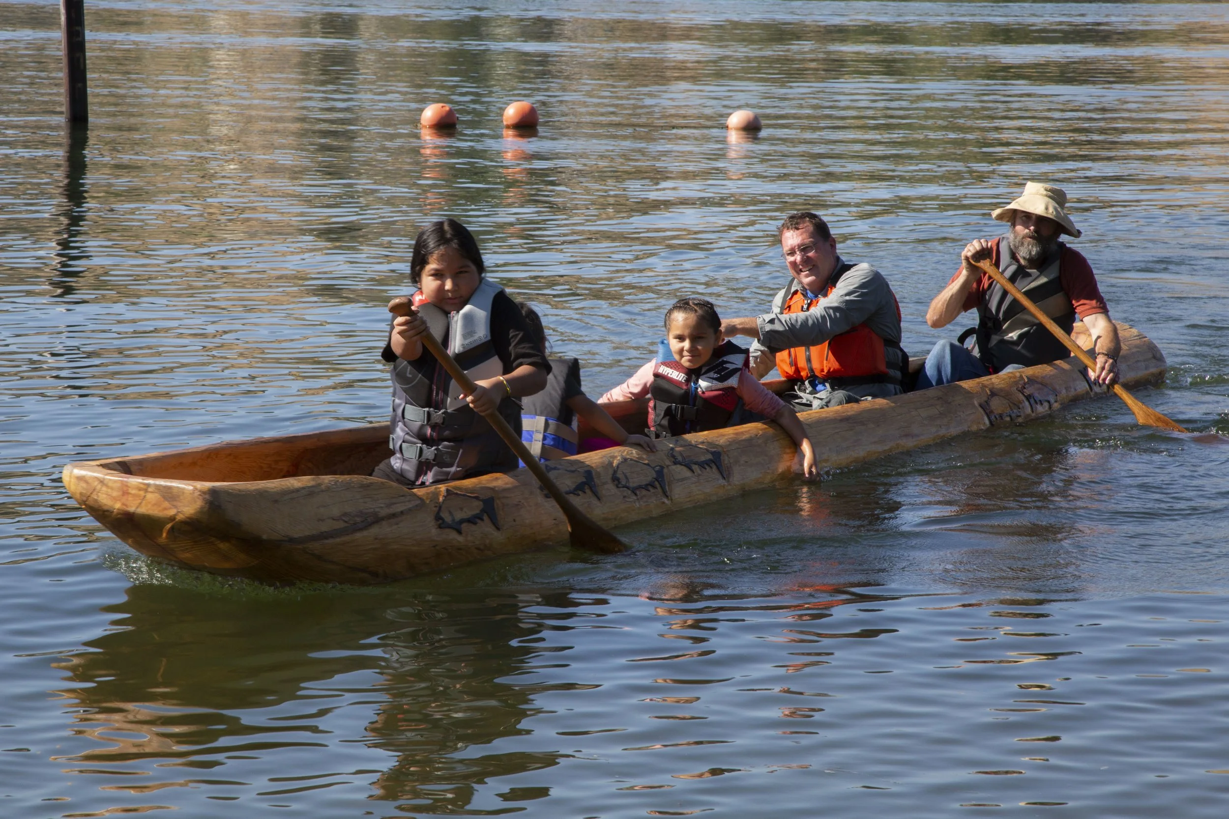 A family of four in a wooden canoe on a calm body of water, with a young girl in the front holding a paddle and a young boy sitting next to her, while two adults paddle at the back. The water has small ripples and orange buoys are floating in the bac