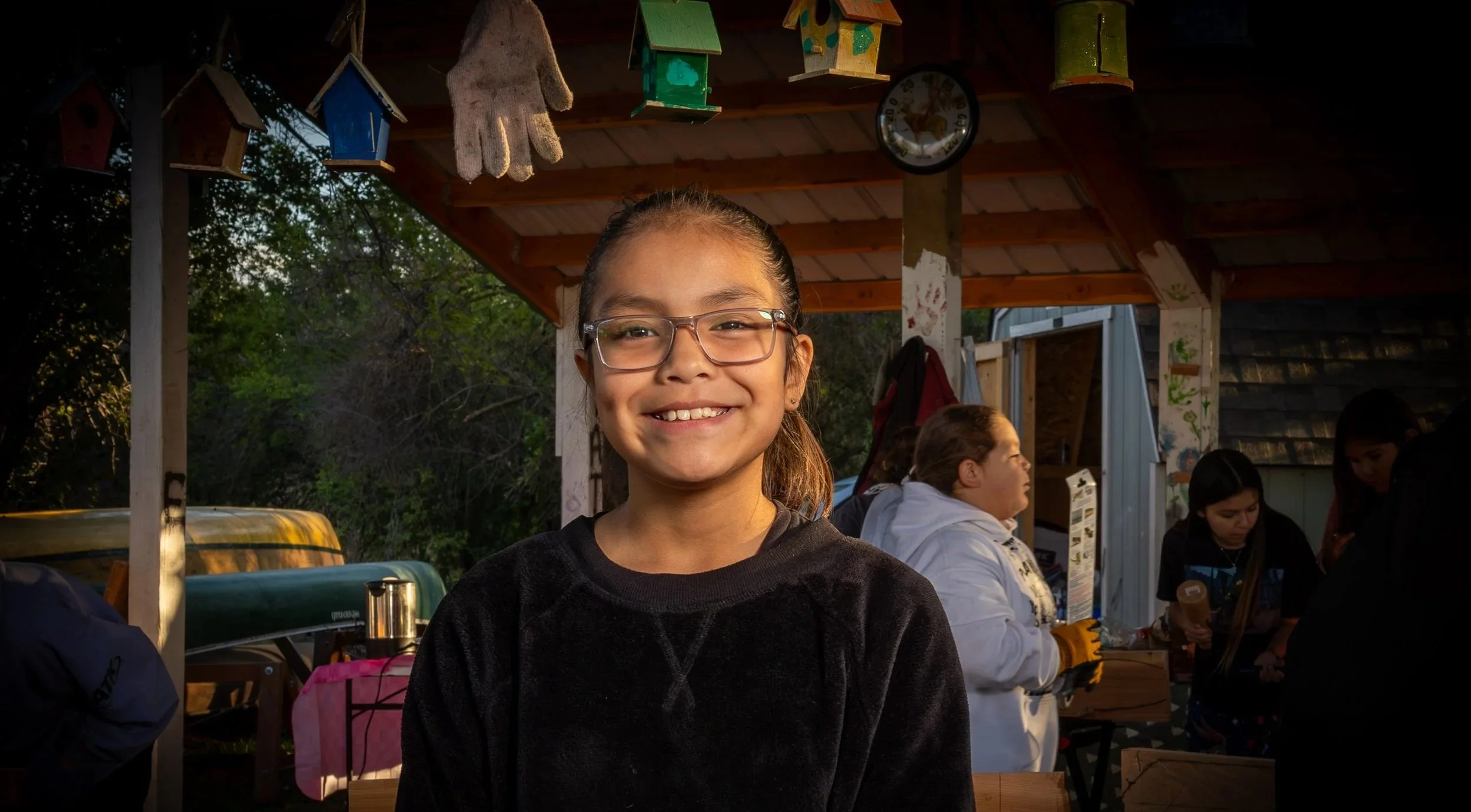 A smiling girl with glasses standing outdoors under a wooden shelter with birdhouses hanging from the ceiling, with other people nearby, some talking and engaging with objects.
