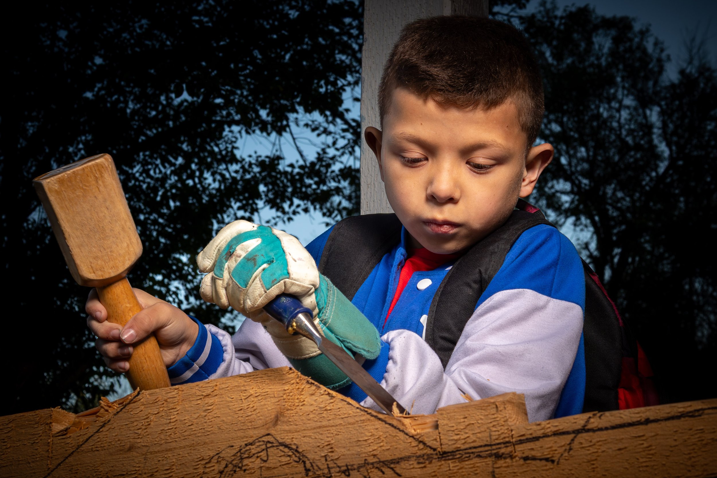 A young boy wearing gloves and a backpack is using a hammer and saw to work on a piece of wood outdoors during the evening, with trees visible in the background.