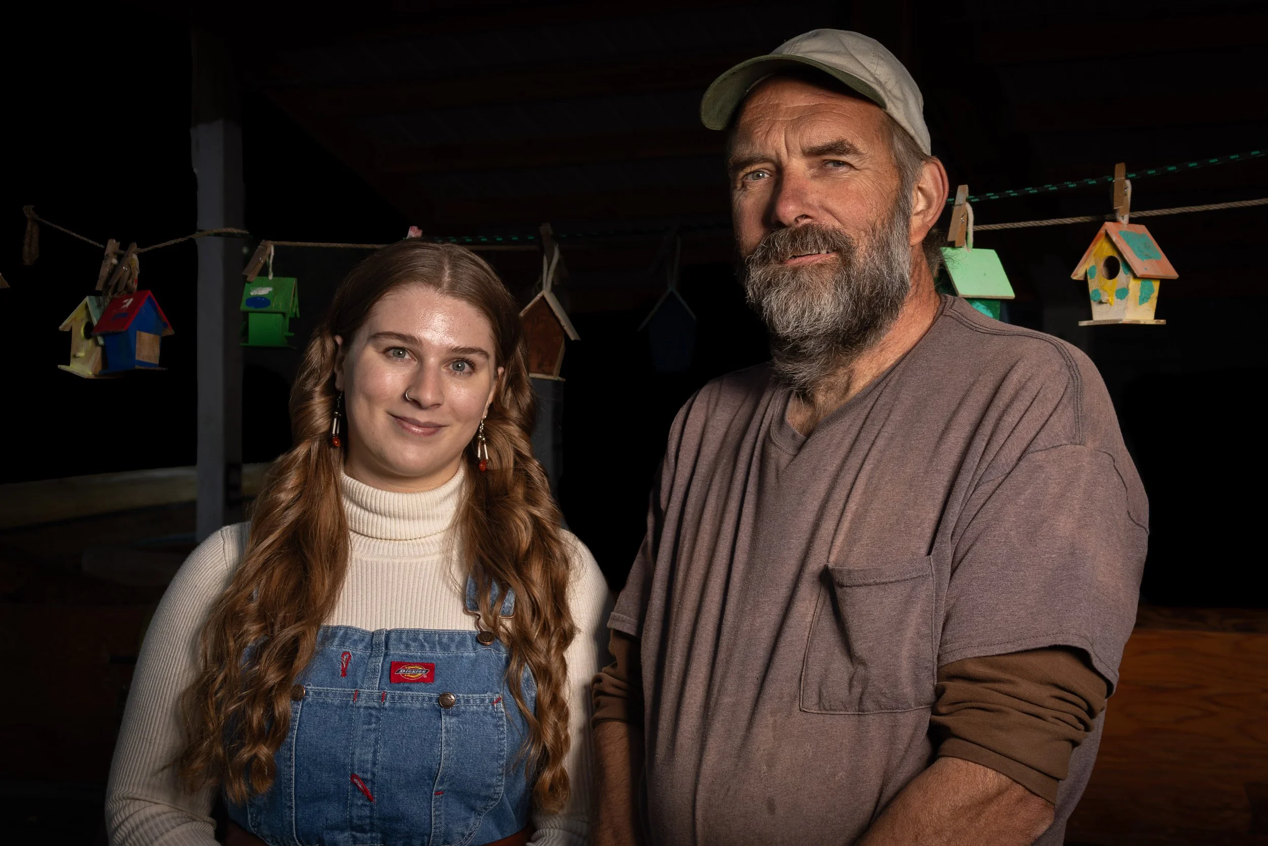 A young woman with long red hair and a man with a beard and light-colored cap standing indoors in front of a string of small colorful birdhouses.
