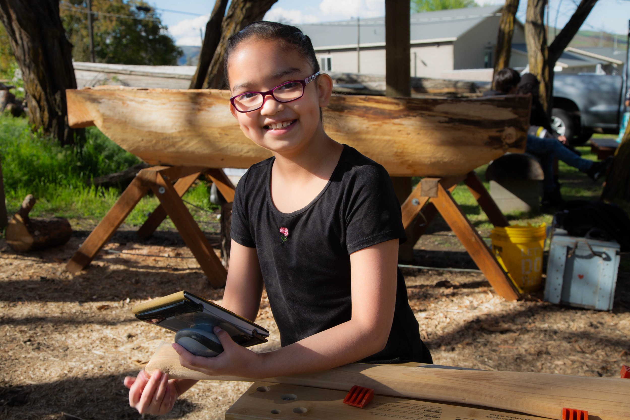 A young girl with glasses smiling while holding a tablet, outdoors during daytime with trees and a wooden structure in background.