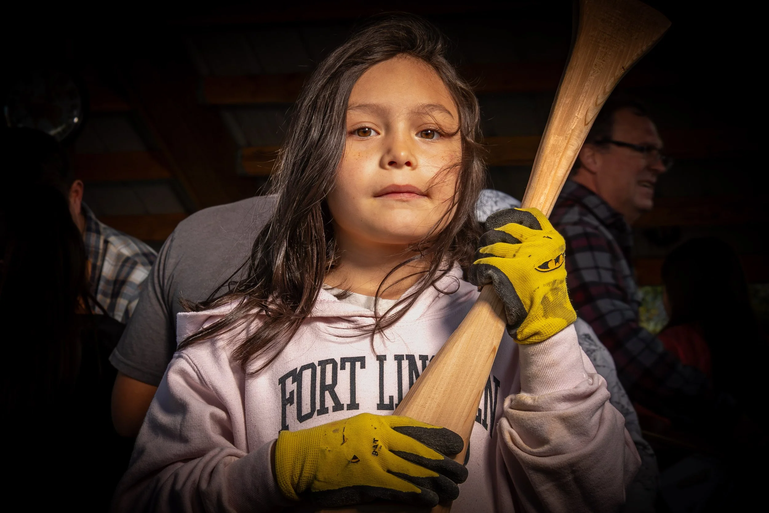 Young girl holding a baseball bat, wearing yellow work gloves and a gray hoodie, with a serious expression. Two men sit in the background, one smiling, in a dimly lit indoor setting.