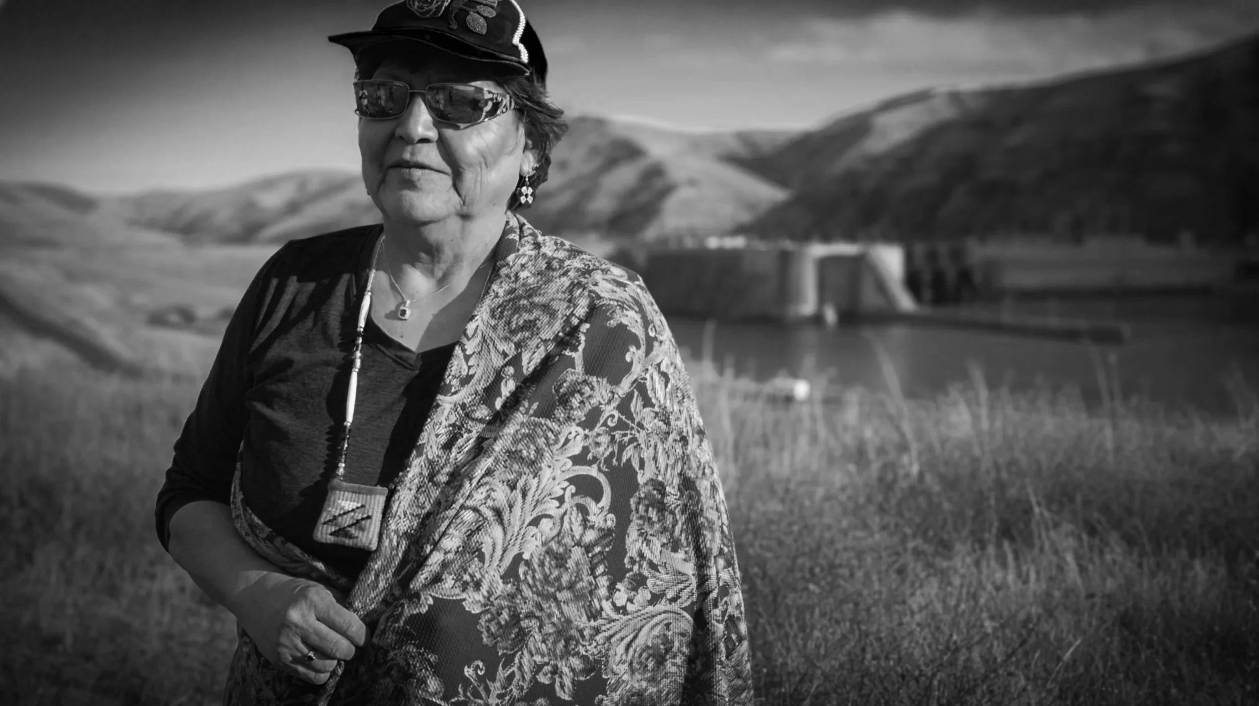 An elderly woman wearing sunglasses, earrings, and a patterned top standing outdoors in front of a rural landscape with hills and a body of water.