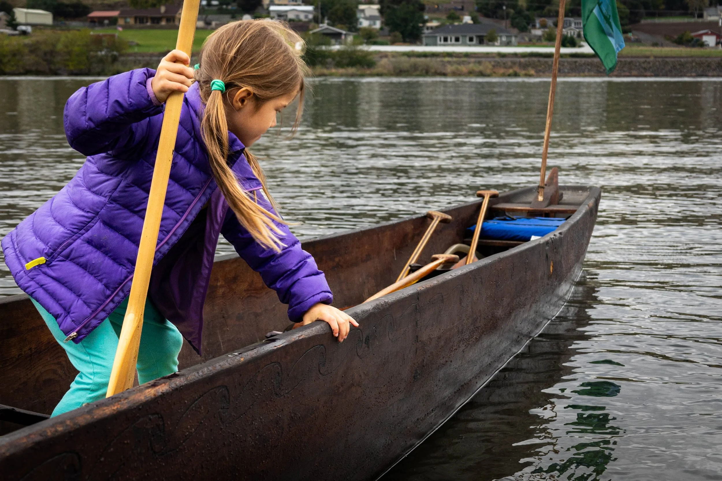 A young girl in a purple jacket and teal pants leaning over the side of a canoe on a river, looking at the water, with houses and trees in the background.
