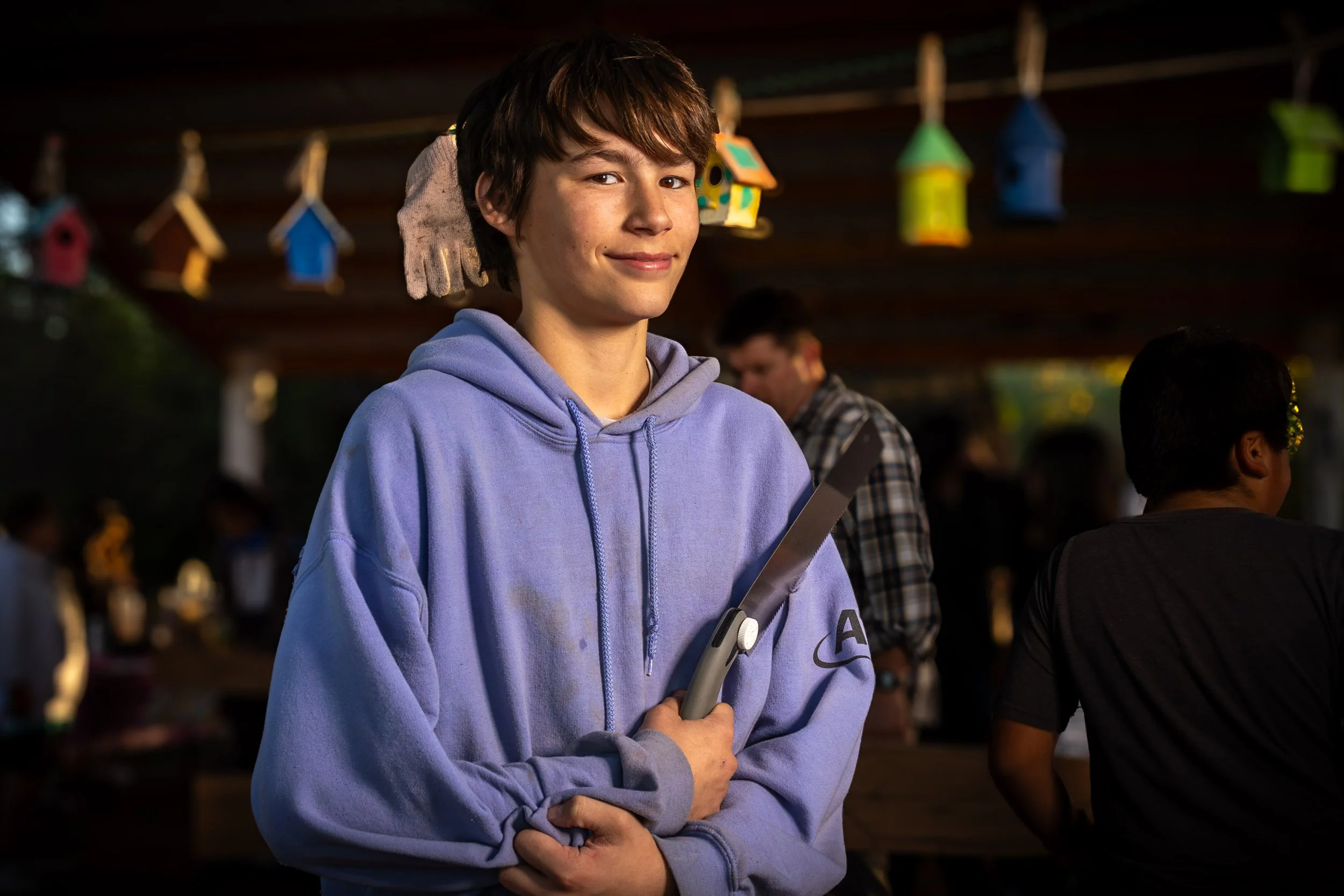Young man with brown hair wearing a purple hoodie, smiling gently, holding a gray object, amid a crowd with colorful hanging birdhouses in the background.