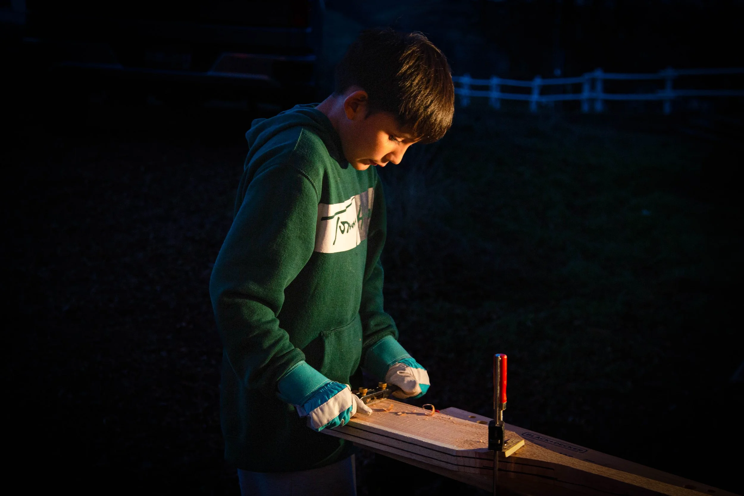 A young boy in a green hoodie working on a science experiment outdoors at night under low lighting, with safety gloves and a small platform, illuminated by a small light source.