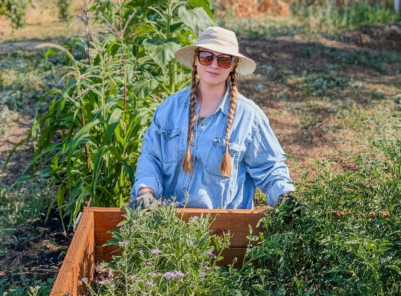 A woman wearing sunglasses, a wide-brimmed hat, a denim shirt, and gardening gloves standing in a garden with various plants and flowering bushes.