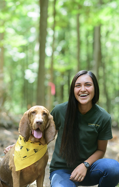 A young woman with long dark hair smiling next to a brown dog wearing a yellow bandana, in a wooded outdoor area.