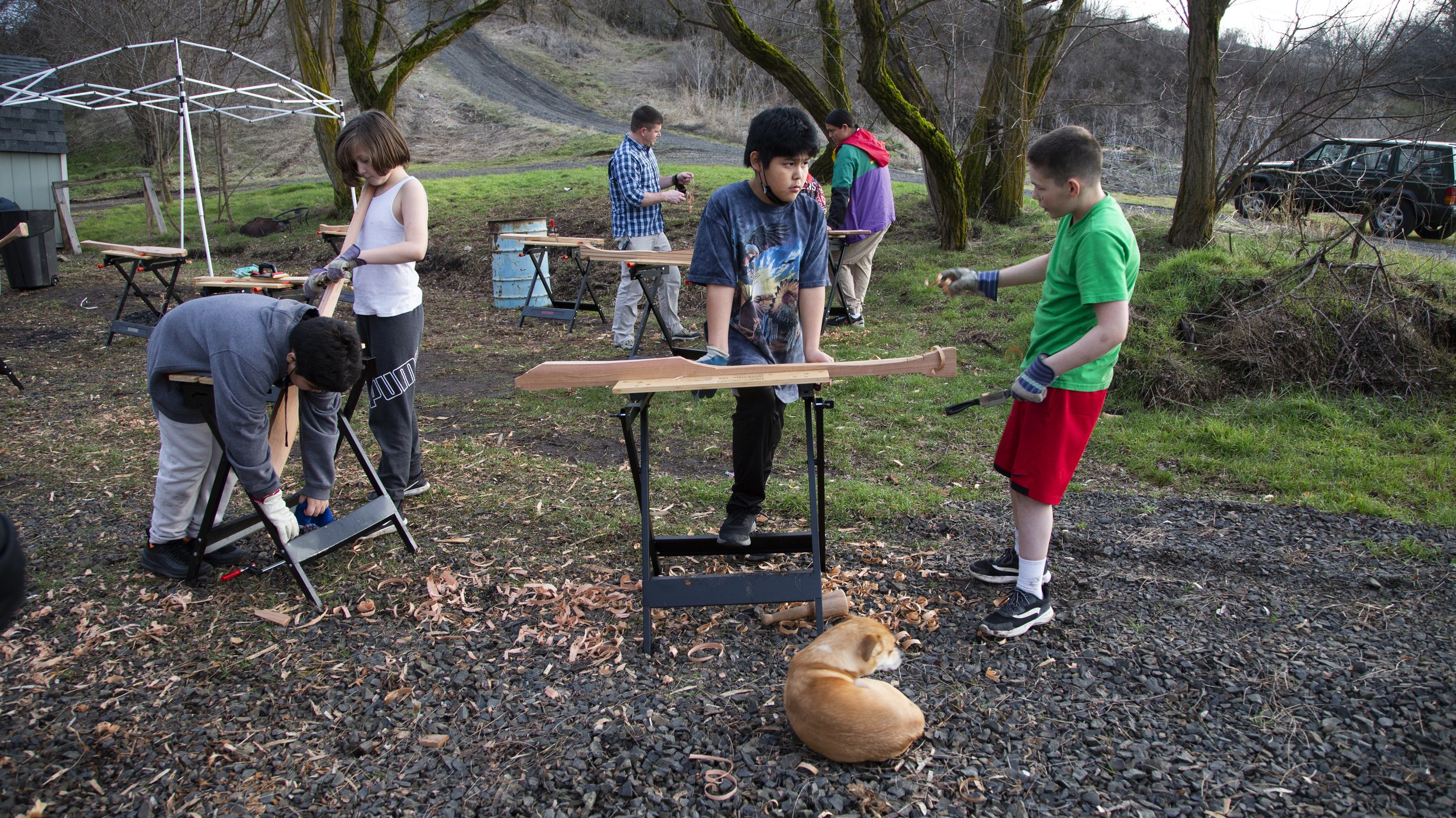 Group of children woodworking outdoors in a yard with trees, working at saw horses and tables with wood pieces, a dog lying on the ground, and a parked vehicle in the background.