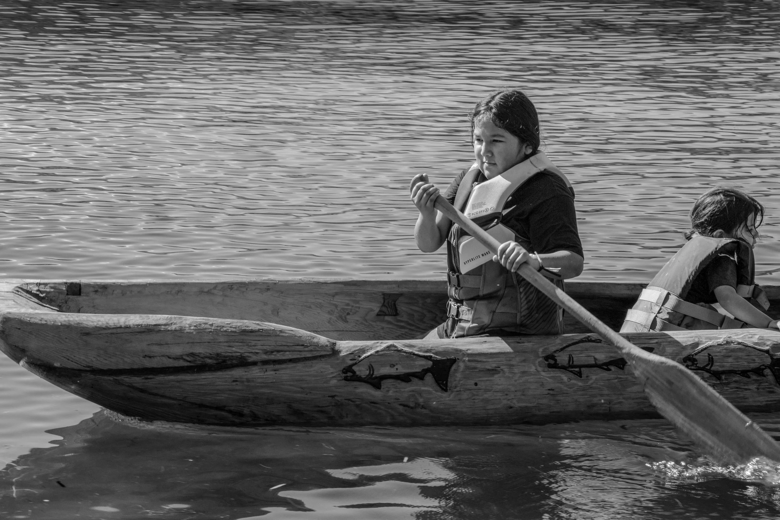 A young girl with wet hair wearing a life jacket paddling a wooden canoe on a calm body of water, with another person partially visible in the canoe.