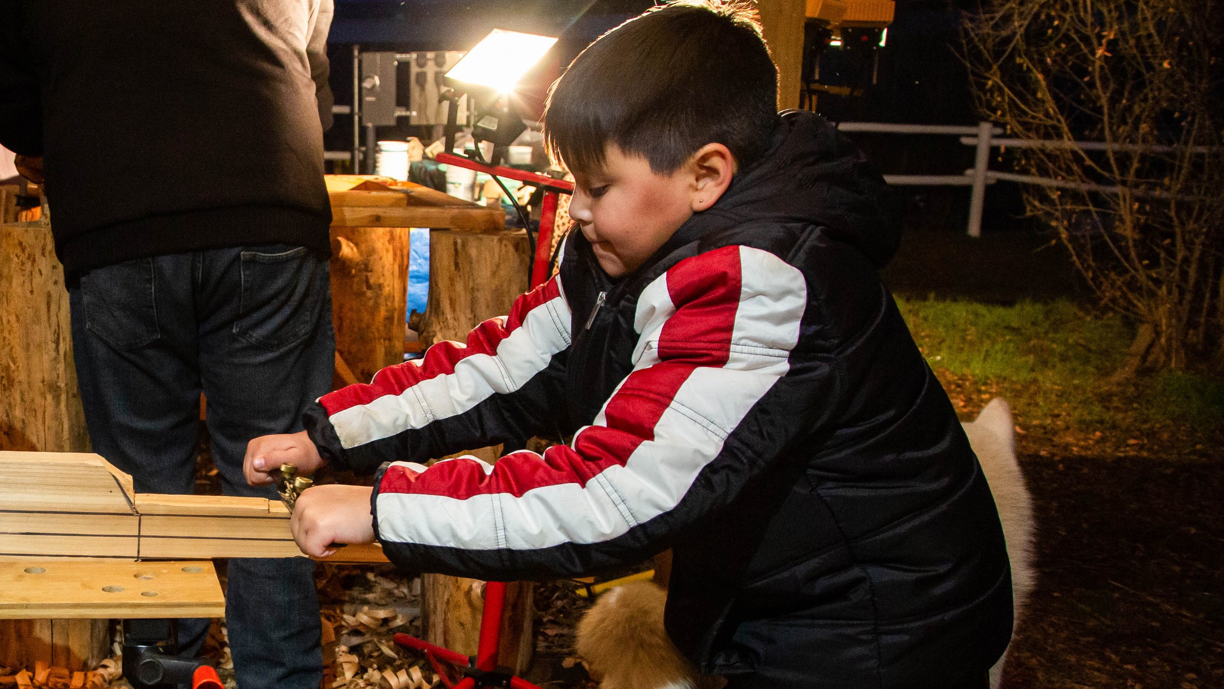 A young boy with dark hair, wearing a black jacket with red and white stripes, is working on a woodworking project at a workbench during an outdoor event at night. There is a bright light and other people around him.