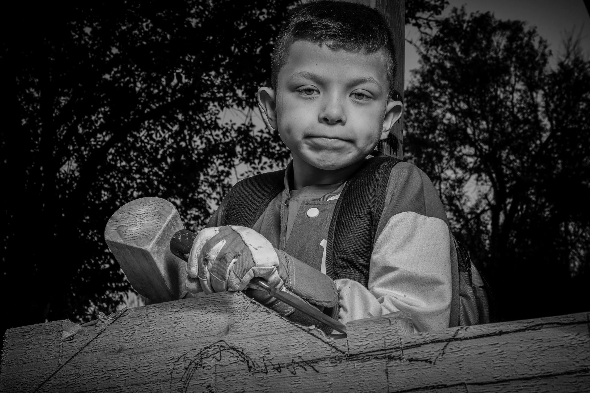 A young boy with short hair, wearing gloves and a backpack, is operating a chainsaw on a large wooden log outdoors. Trees and a cloudy sky are visible in the background.