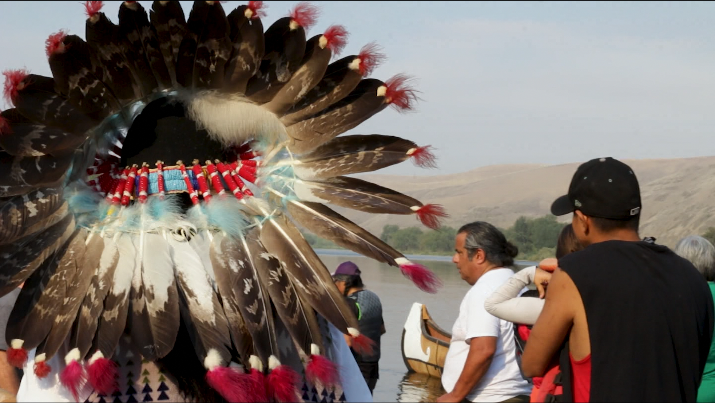 A person wearing a traditional Native American headdress with brown, white, and red feathers, and beadwork, is accompanied by a group of people near a body of water and hills in the background.