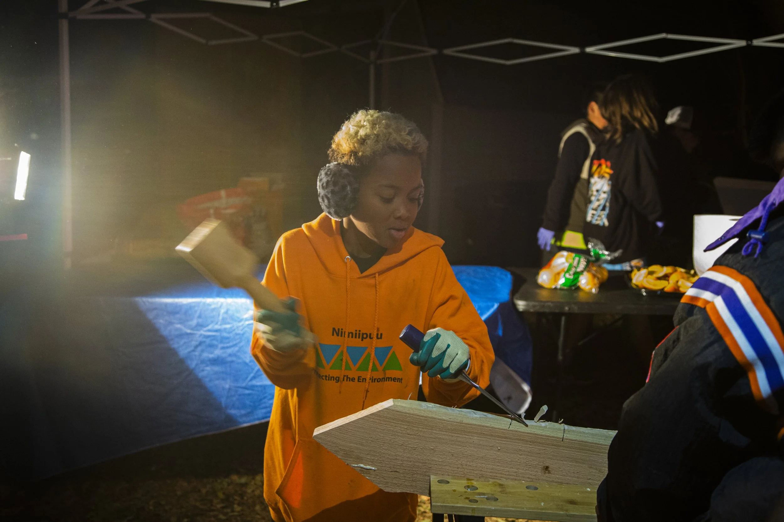 A woman in an orange hoodie working on a wooden project at an outdoor night event, with several other people in the background under canopy tents.