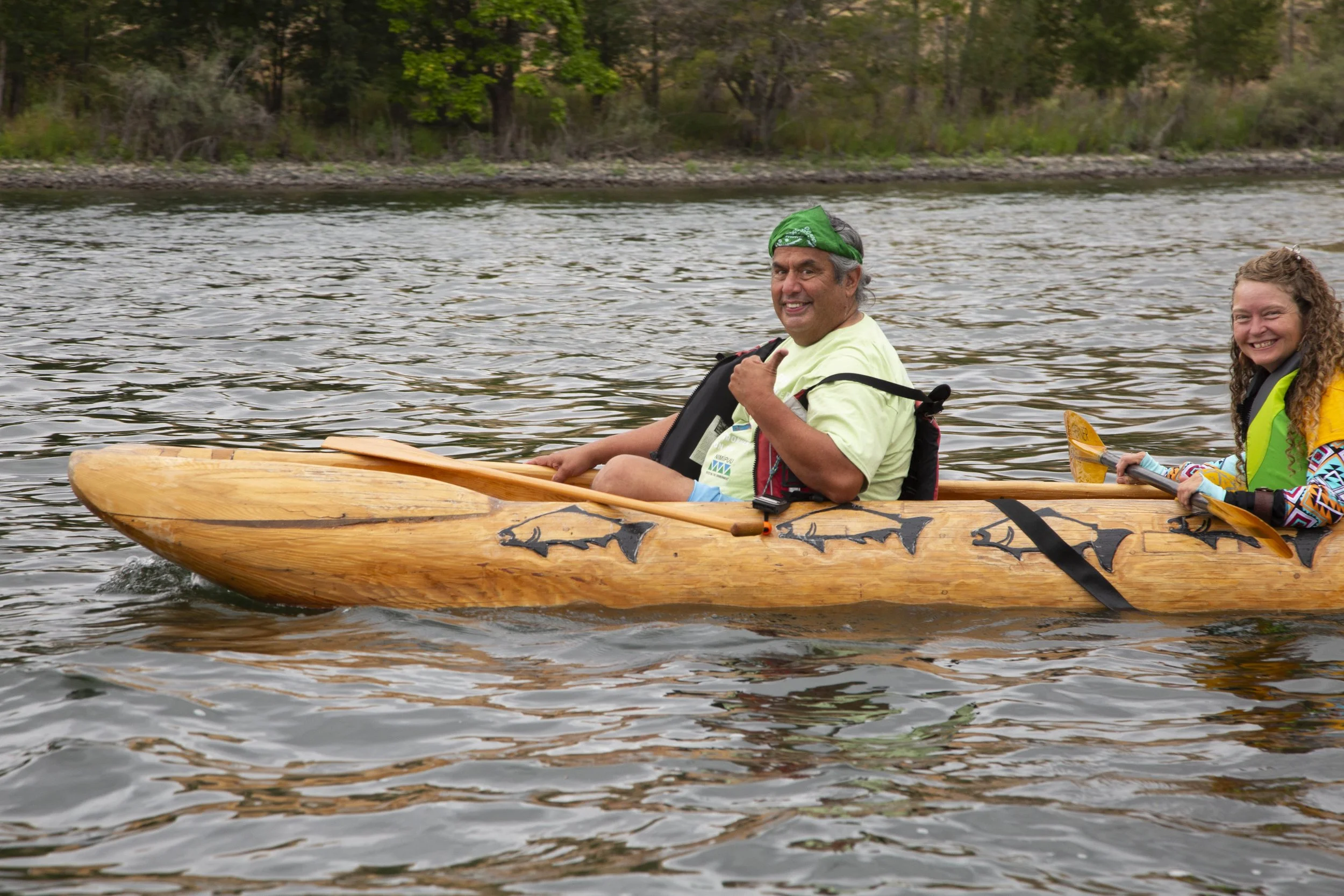 Two smiling people are sitting in a wooden canoe on a river, surrounded by trees.