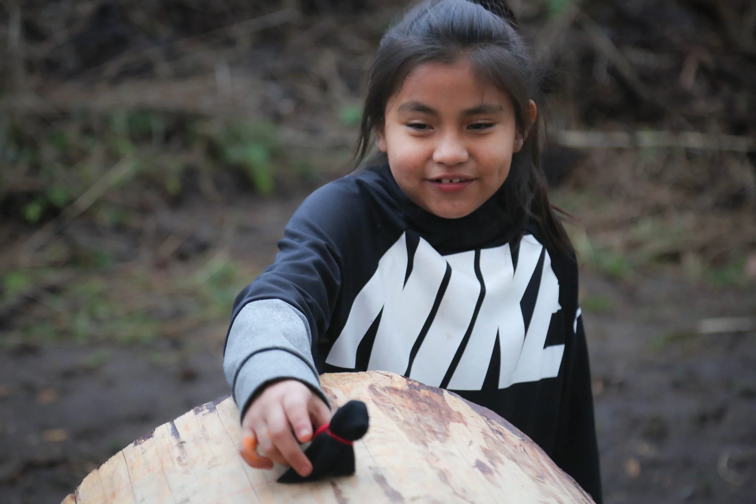 A young girl with long dark hair, smiling, wearing a black and white striped jacket and black gloves, is sitting outdoors on a log or a piece of wood, holding a saw, with a blurred natural background of trees and grass.