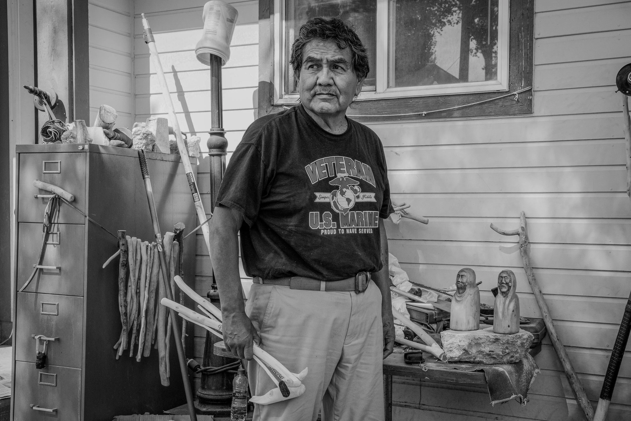 Black and white photo of a middle-aged man standing on a porch, surrounded by various objects including sculptures, tools, and furniture. He is wearing a T-shirt with a graphic and text, and light-colored pants. The background shows a window and laye