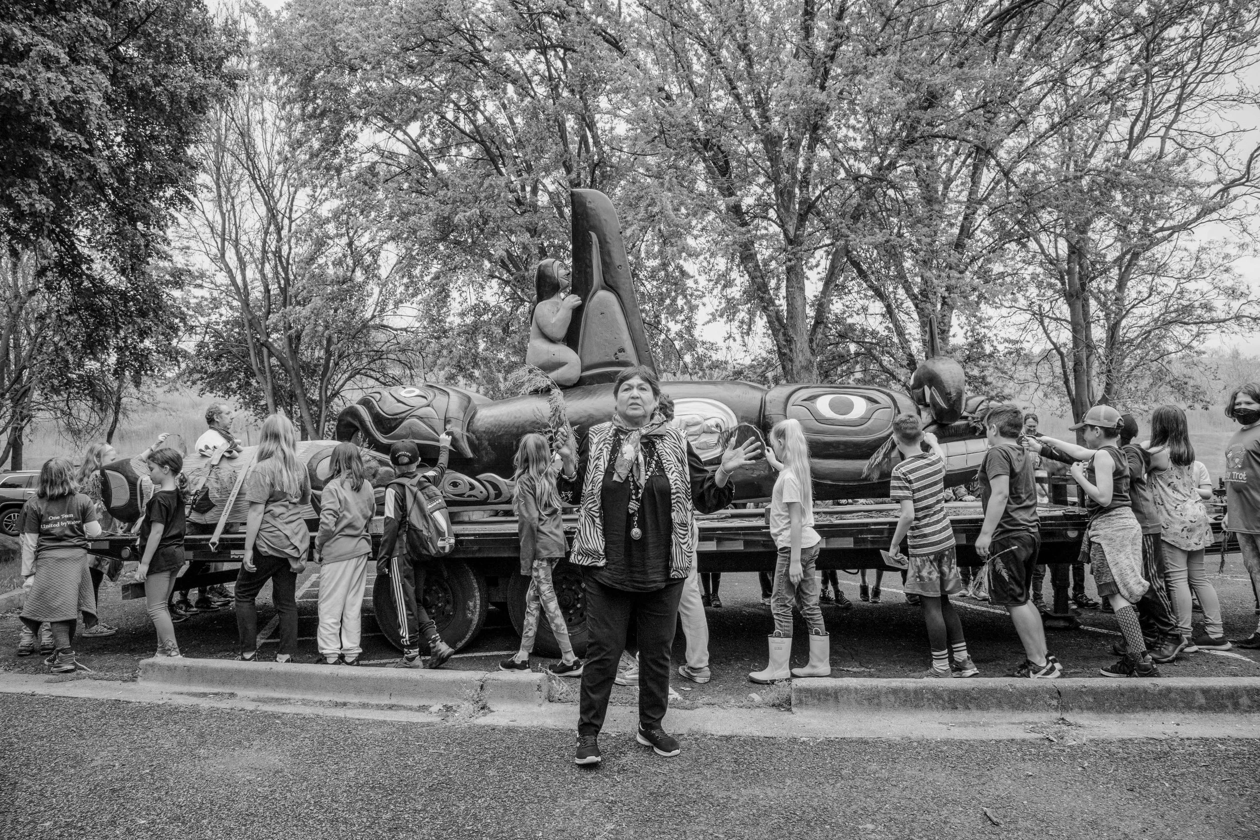 A black-and-white photo of a woman standing in front of a decorated float with children around her. The float features a large figure and an animal, possibly a goose, with tall trees in the background.