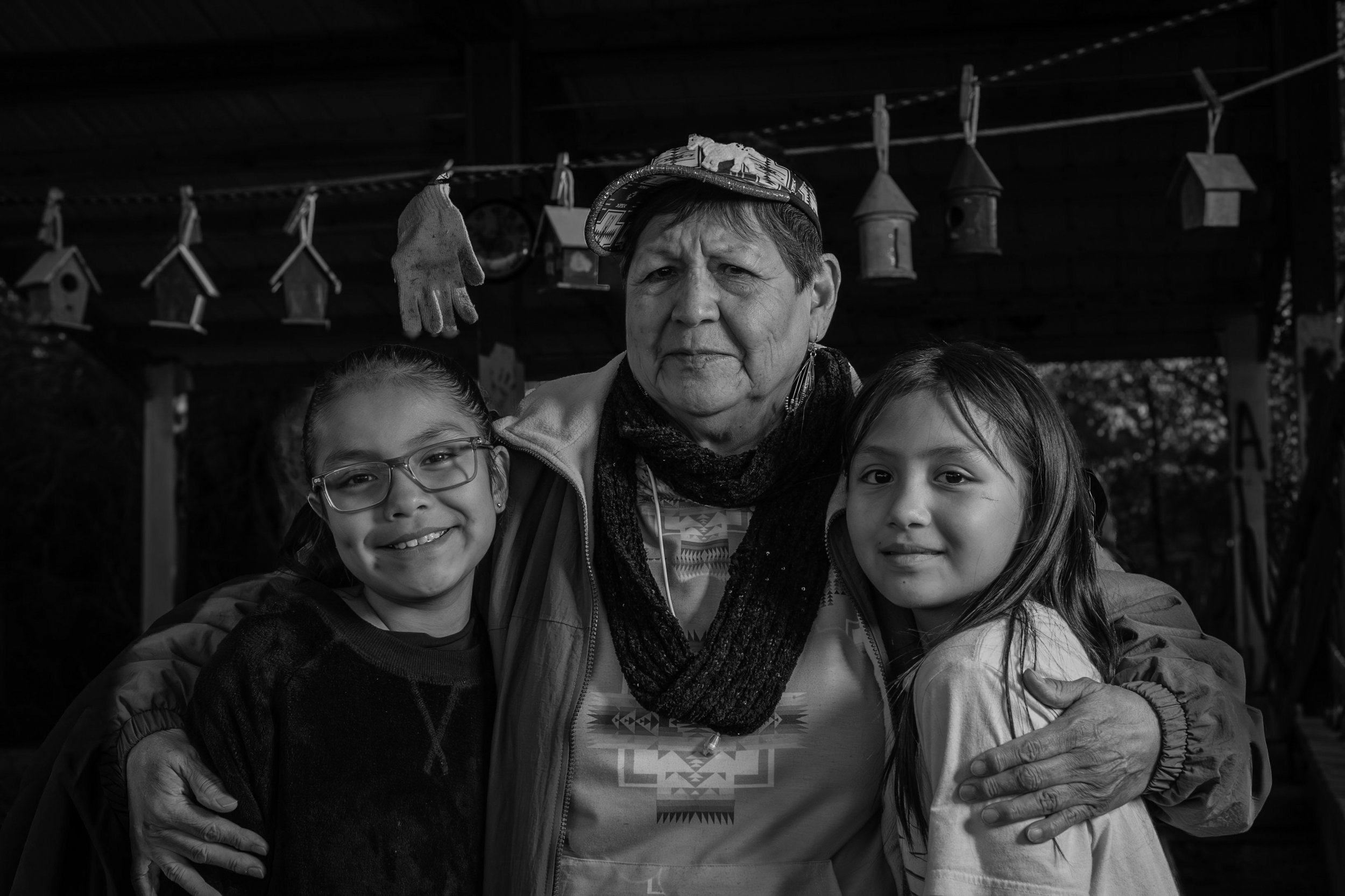 Black and white photo of an older woman with short hair wearing a patterned shirt, a scarf, and a headscarf, standing outdoors with two young girls, one with glasses and the other with long hair, in front of a wooden structure with hanging birdhouses and gloves. The women and girls are smiling and hugging each other.