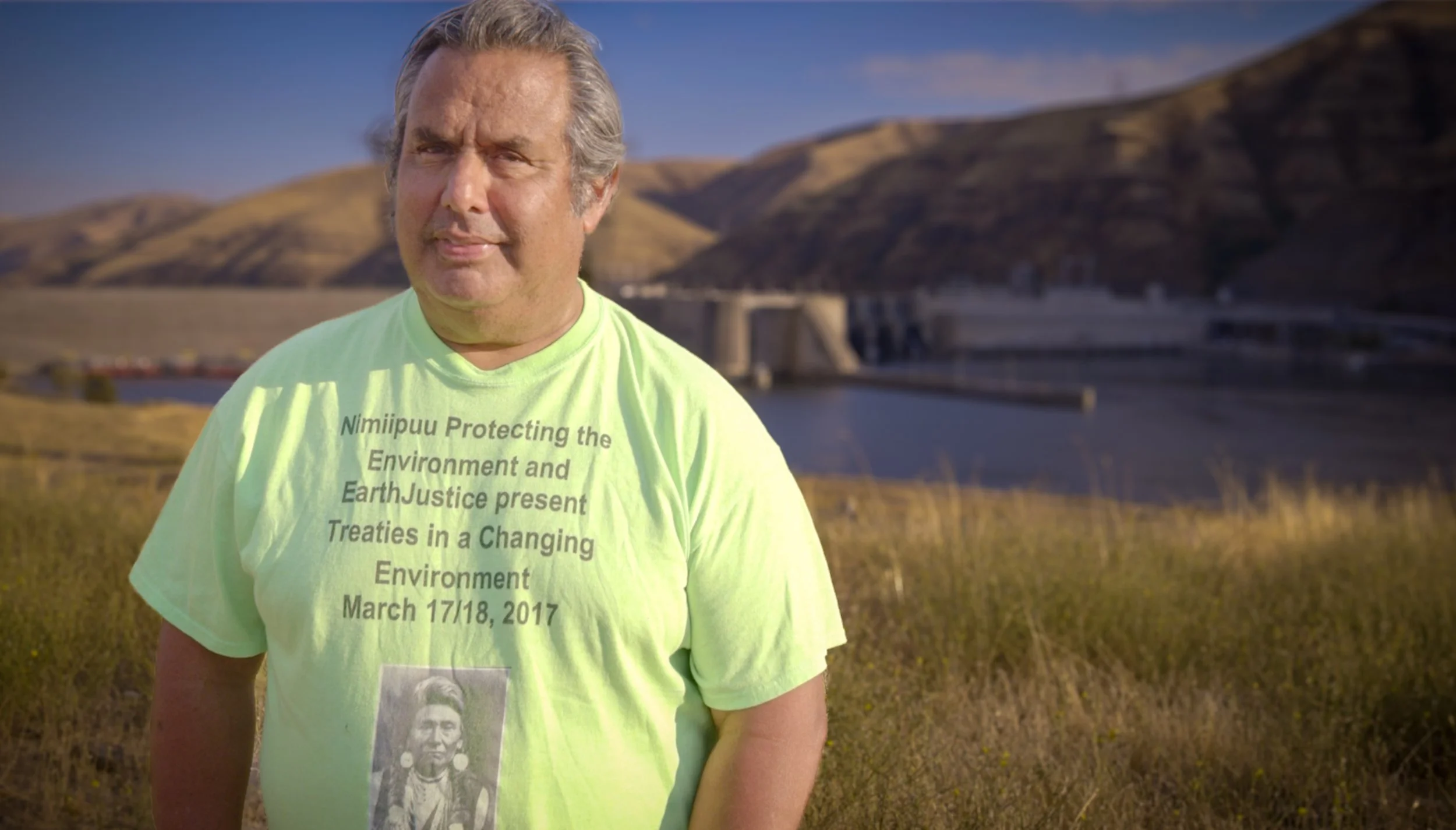 A middle-aged man standing outdoors near a dam and rolling hills, wearing a neon green t-shirt with text about protecting the environment and Earth justice, dated March 17/18, 2017.