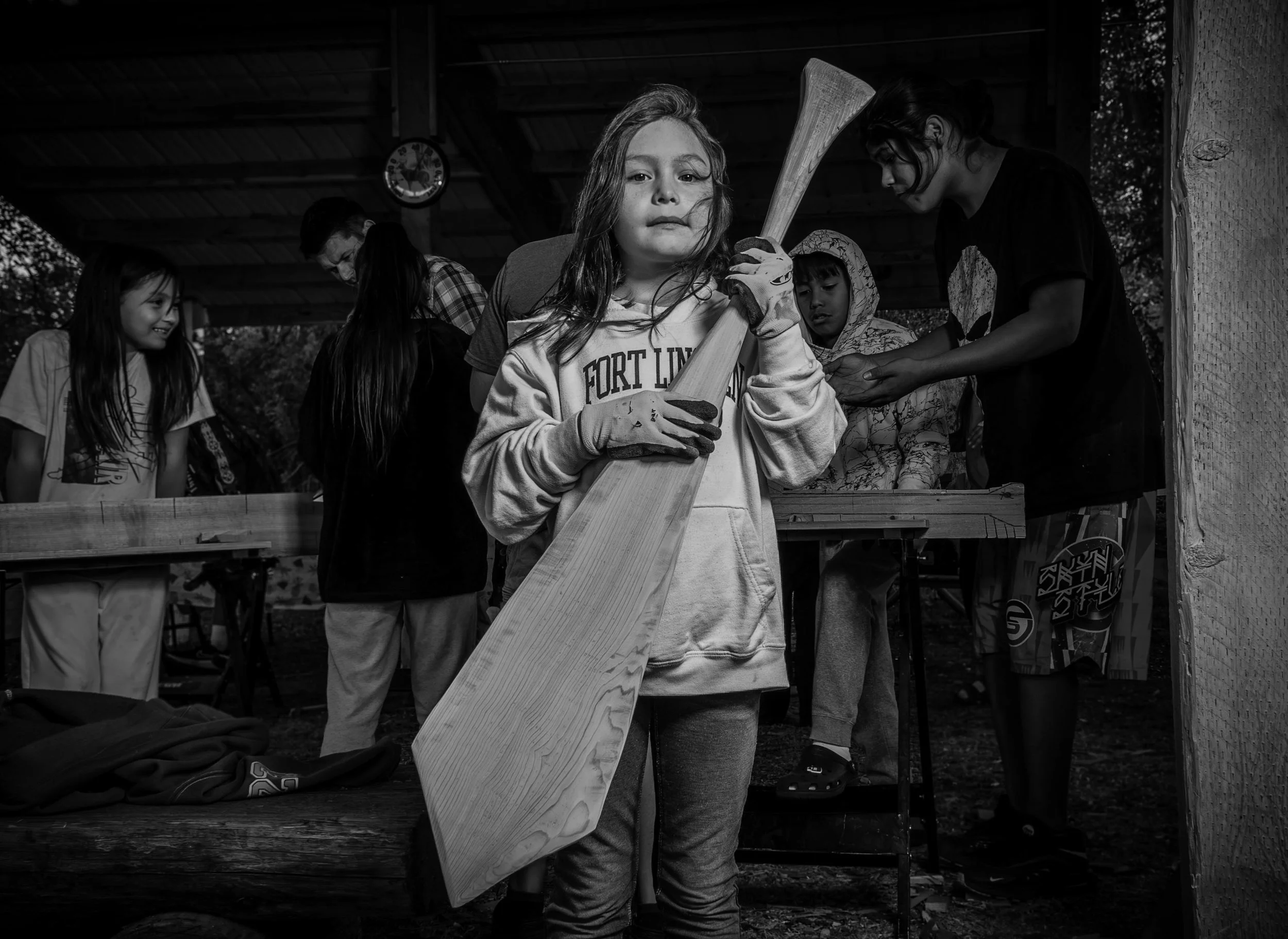 A young girl holding a wooden paddle, surrounded by other children and adults, in a covered outdoor setting, possibly a woodworking or craft project.