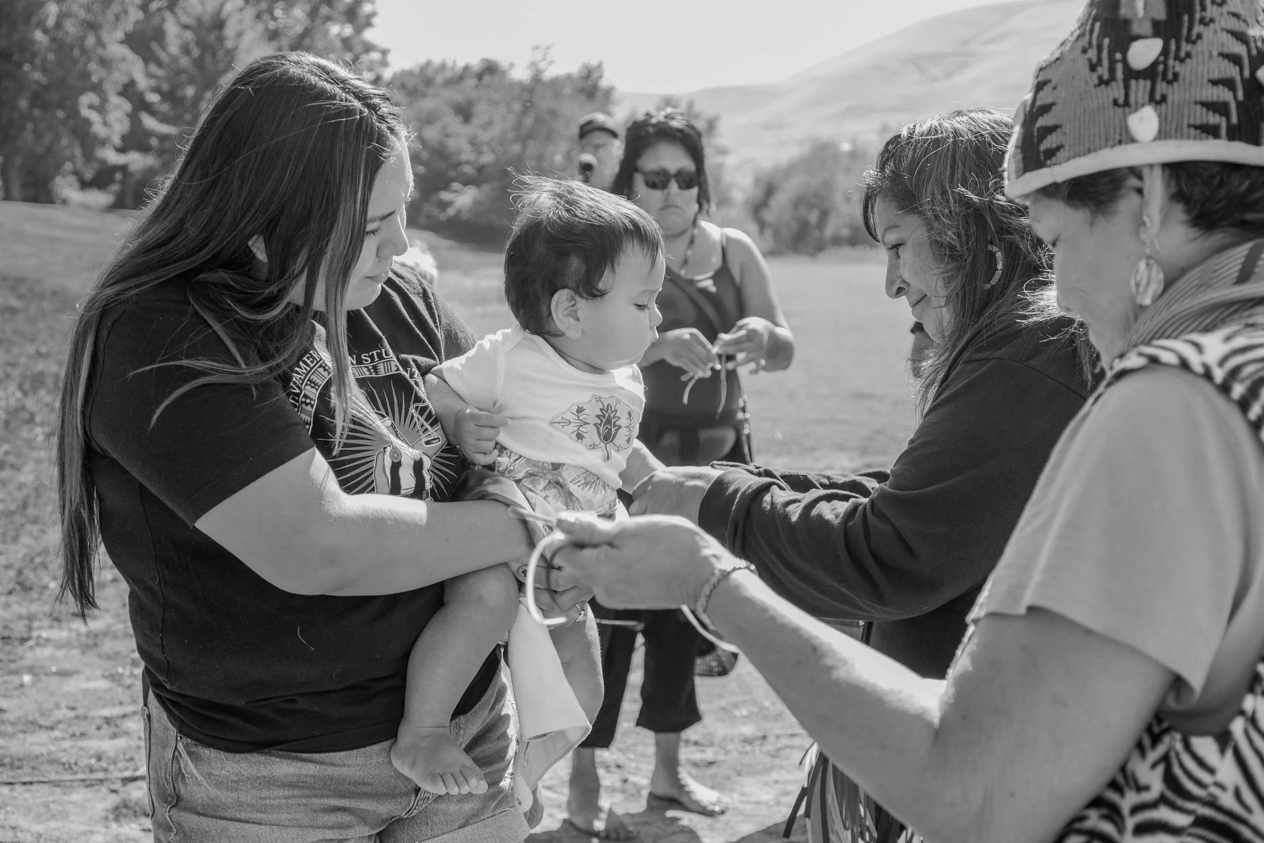 A group of people outdoors, with focus on a woman holding a young child while other adults watch and interact in the background.