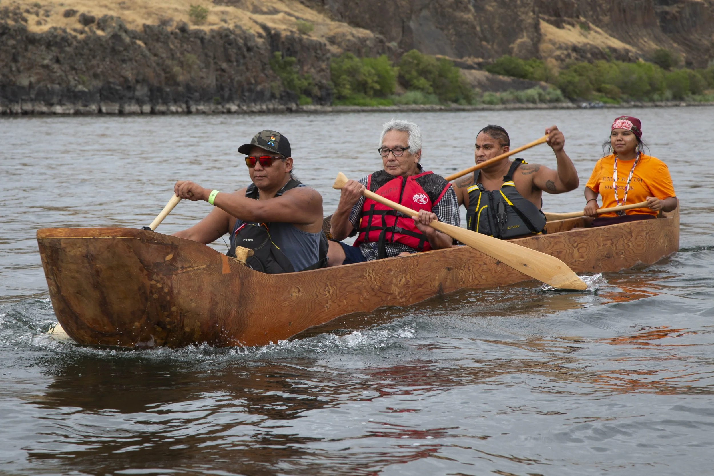 Four people paddle a wooden canoe on a river, with rocky cliffs and green trees in the background.