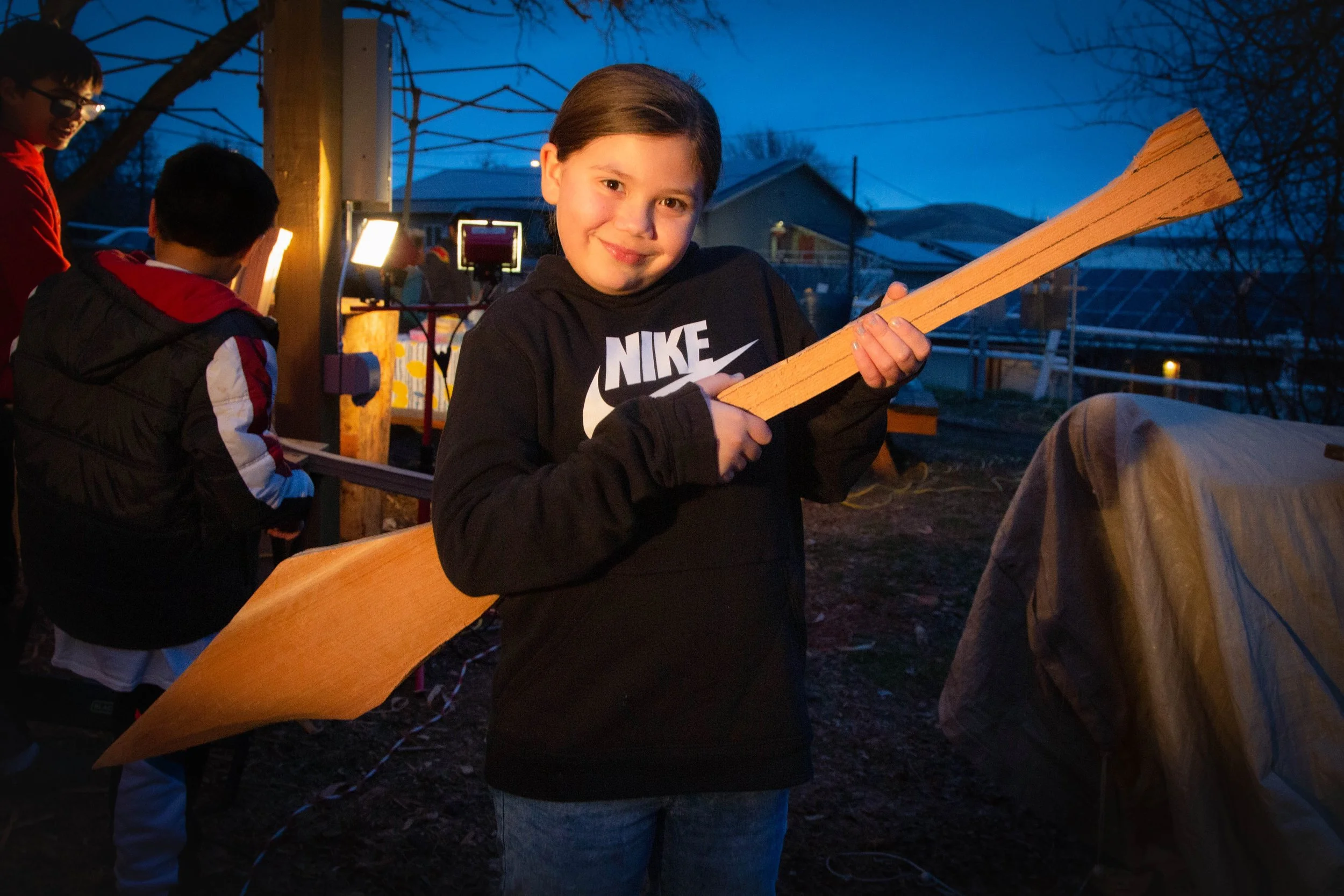 A young girl with dark hair holding a wooden piece, smiling, with other children and outdoor structures in the background during dusk.