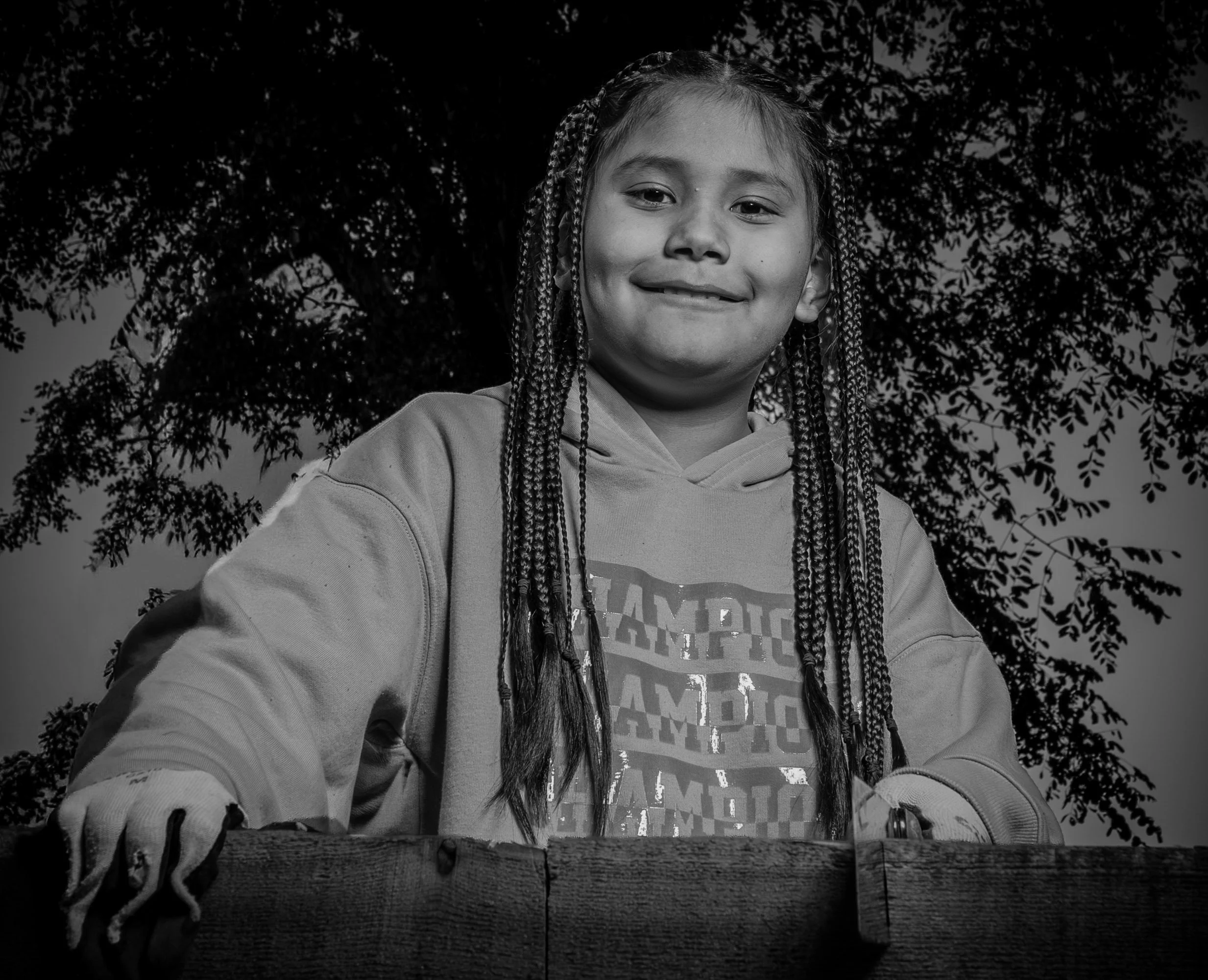 Black and white photo of a young girl with long braided hair, smiling slightly, sitting behind a wooden surface outdoors with trees in the background.