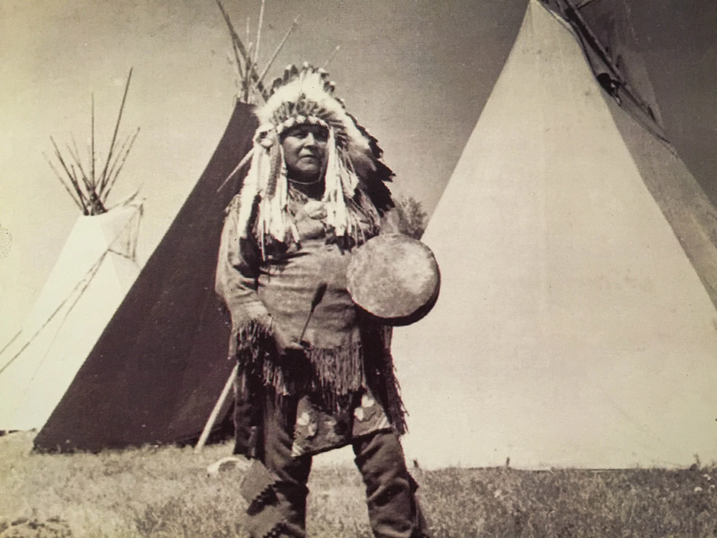 A Native American person wearing a traditional feather headdress and holding a drum, standing outdoors at night.