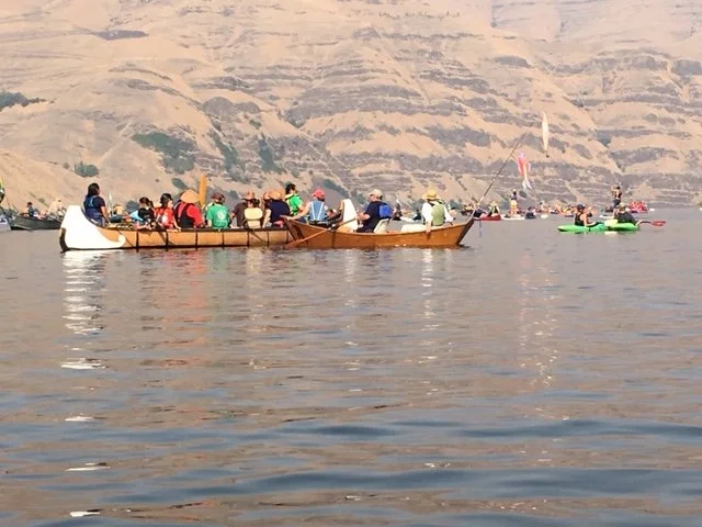 Several people are in small traditional wooden boats on calm water with a mountainous landscape in the background.