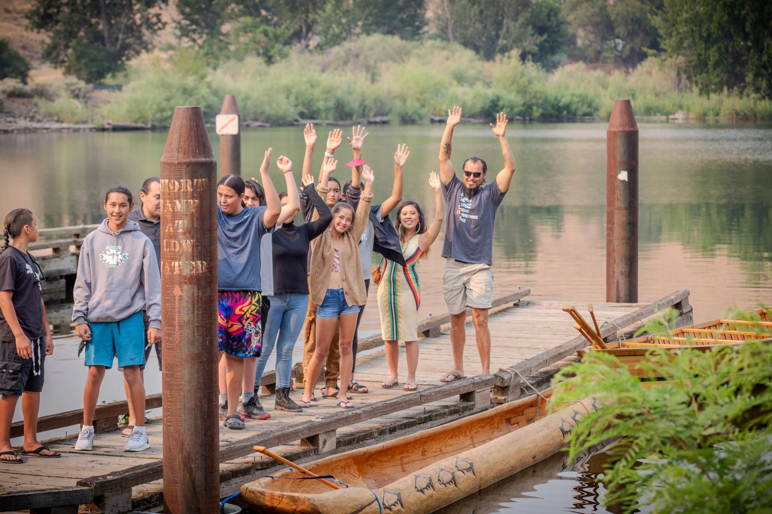 A group of people standing on a wooden dock by the water, waving and smiling, with trees and nature in the background.