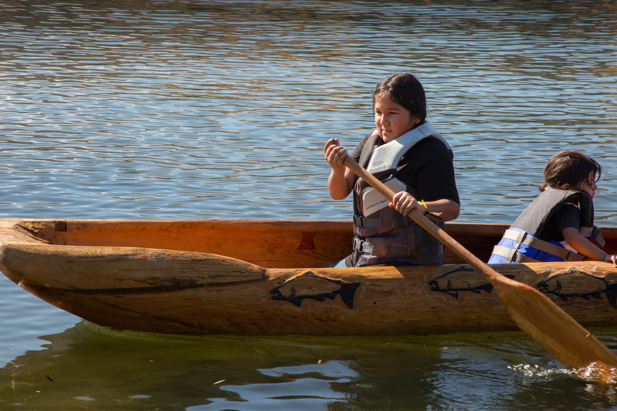 Two children in life jackets paddling a wooden canoe on a body of water.