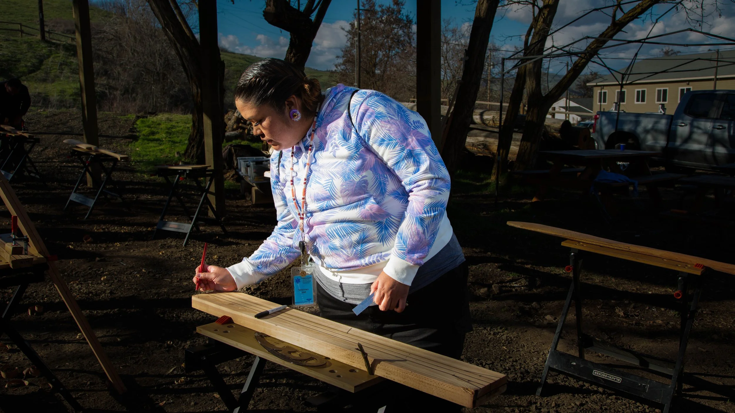 Woman wearing a tropical-patterned shirt and lanyard working on a woodworking project outdoors with various tables, tools, and trees in the background.