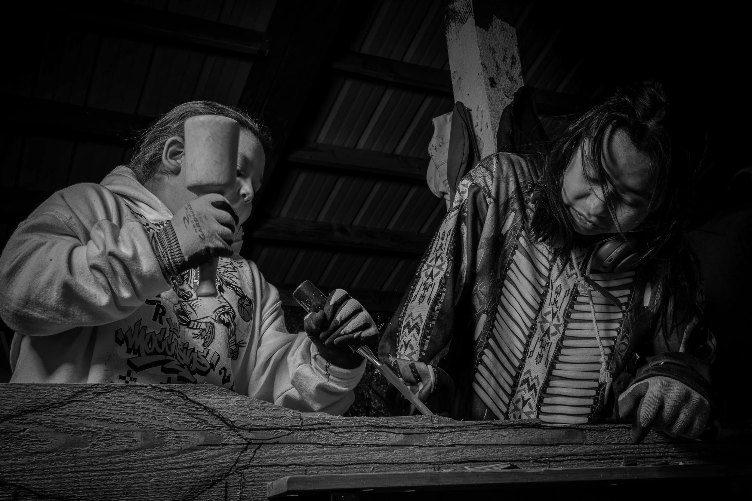 Two women arm wrestling, one holding a small hammer, in a rustic setting with wooden walls.