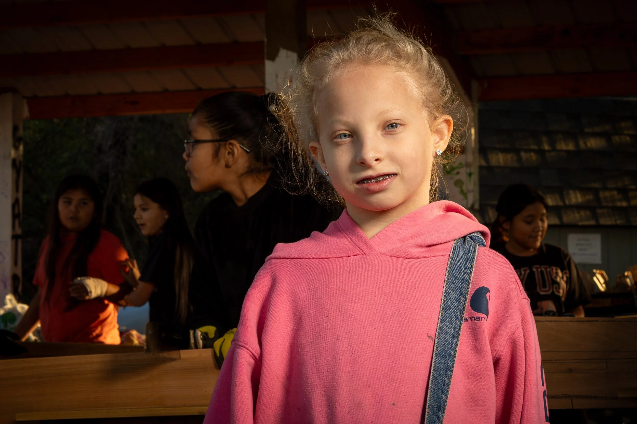A young girl with blonde hair in a ponytail wearing a pink hoodie with a denim strap over her shoulder, standing outdoors with a group of children in the background, some with dark hair and two wearing glasses, under a shelter with a wooden roof.