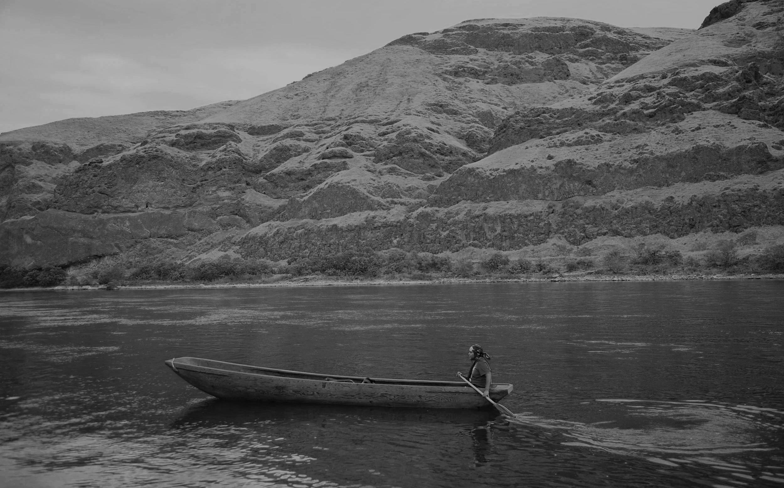 A person in a small boat on a body of water, with a mountain or hillside in the background. The person appears to be sitting and paddling with a long oar.