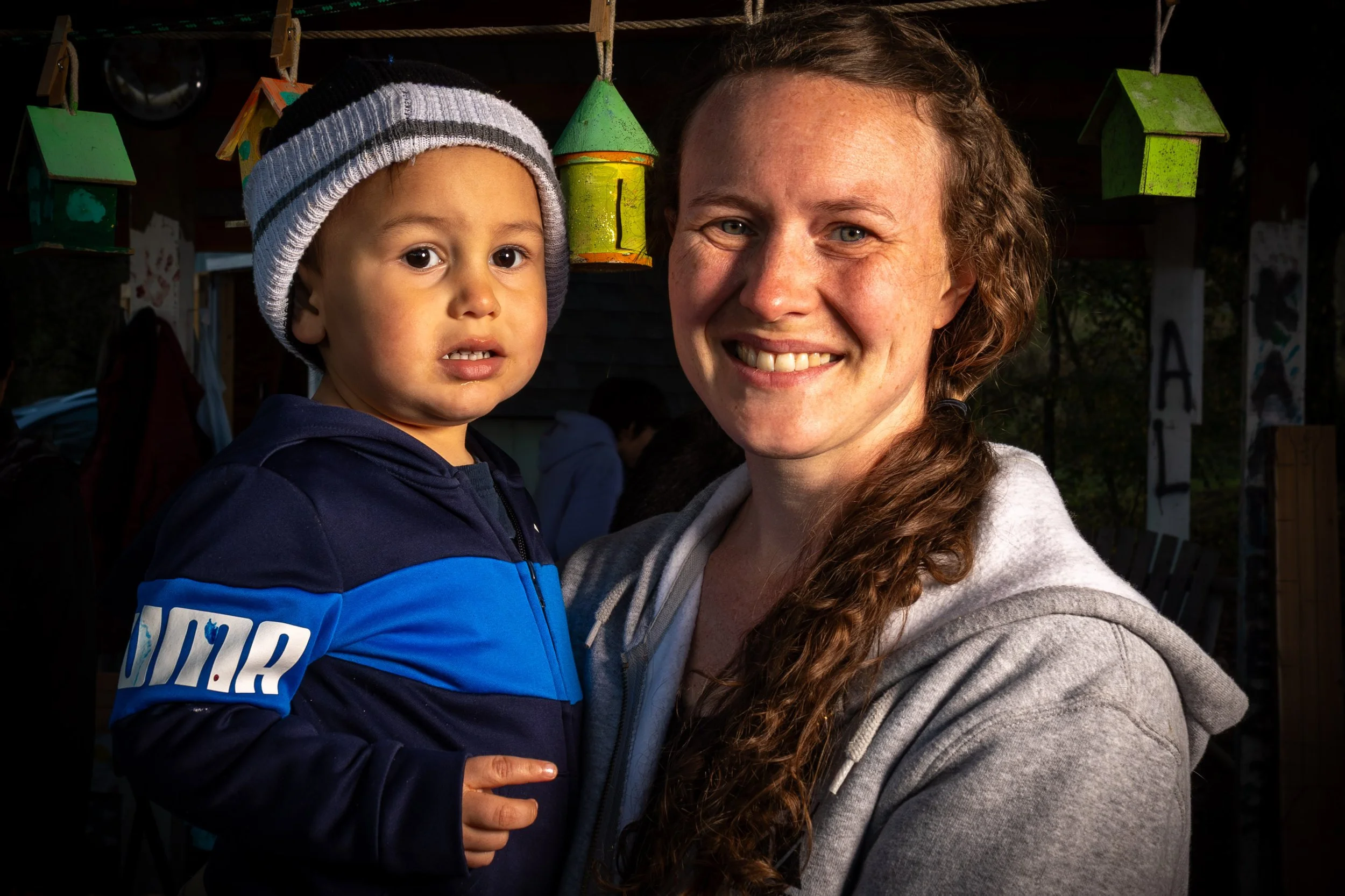 A woman with curly hair smiling while holding a young boy with a striped beanie hat and a blue jacket, both looking at the camera.