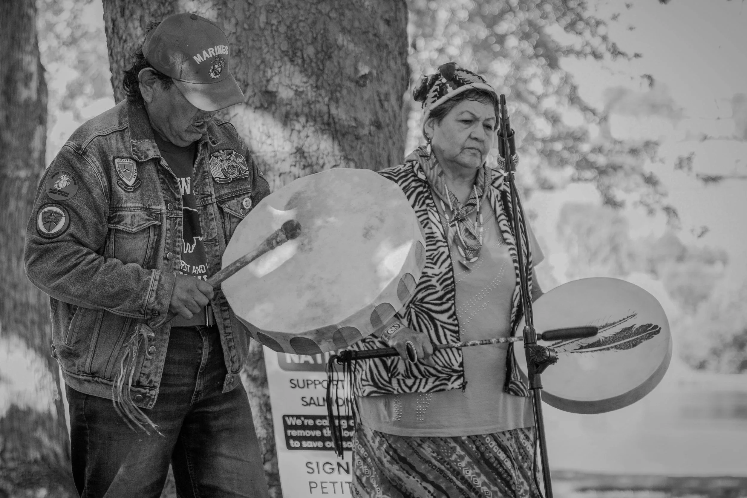 A man and a woman stand outdoors near a tree. The man is wearing a denim jacket and cap with Marine patches, looking down and holding a shovel. The woman, wearing a patterned headscarf and blouse, appears somber and is holding a large circular object