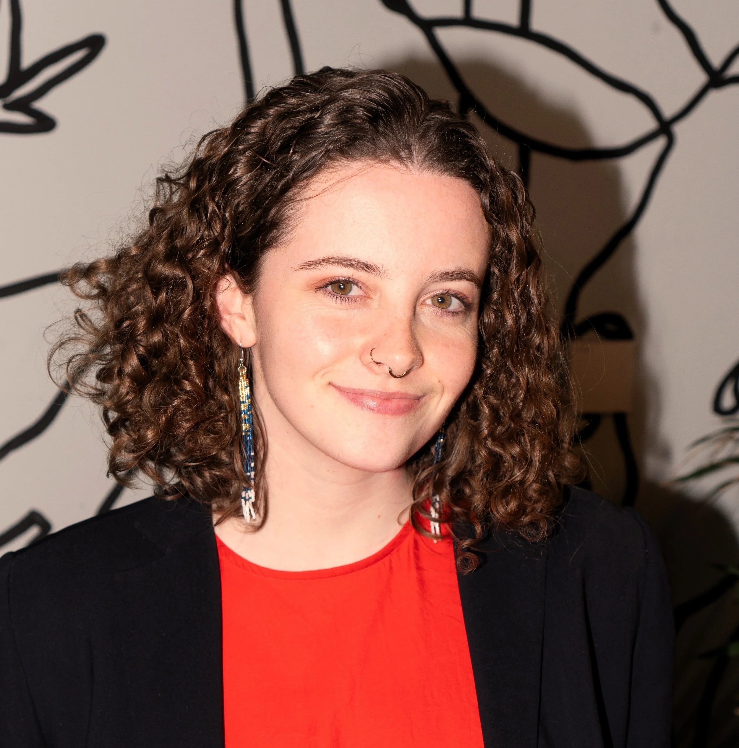 Close-up of a woman's face with curly brown hair, wearing earrings, a nose ring, and a black blazer over a red top, standing in front of a black and white abstract background.