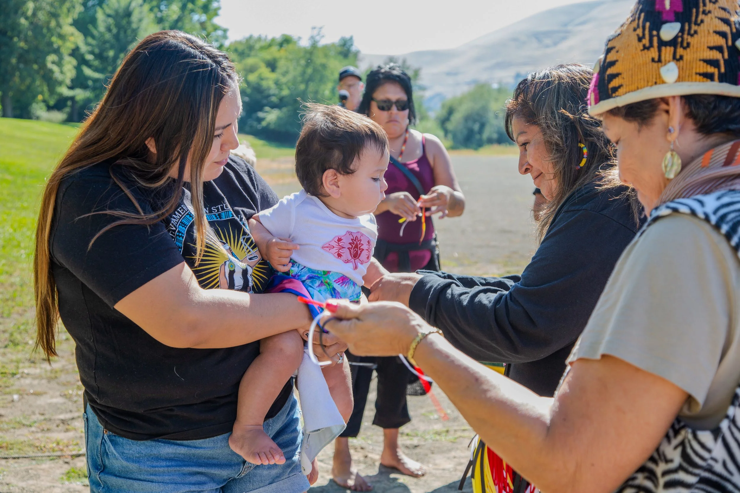 Group of Indigenous women and children celebrating a ceremony outdoors during the day, with mountains and trees in the background.
