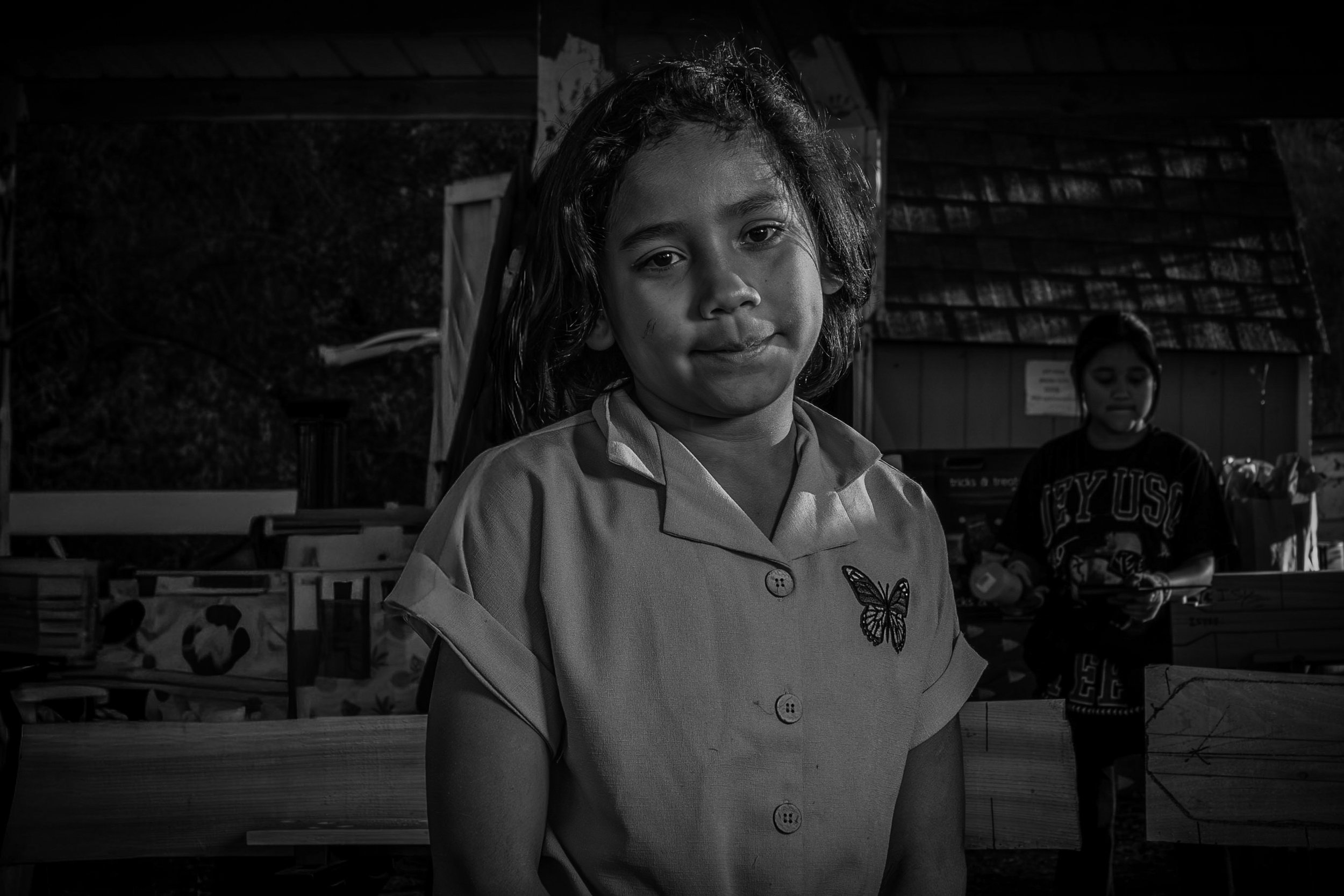 A young girl with wet hair standing outdoors with a subtle smile. She is wearing a button-up shirt with a butterfly design. In the background, another girl in a dark shirt is visible, along with wooden structures and trees.