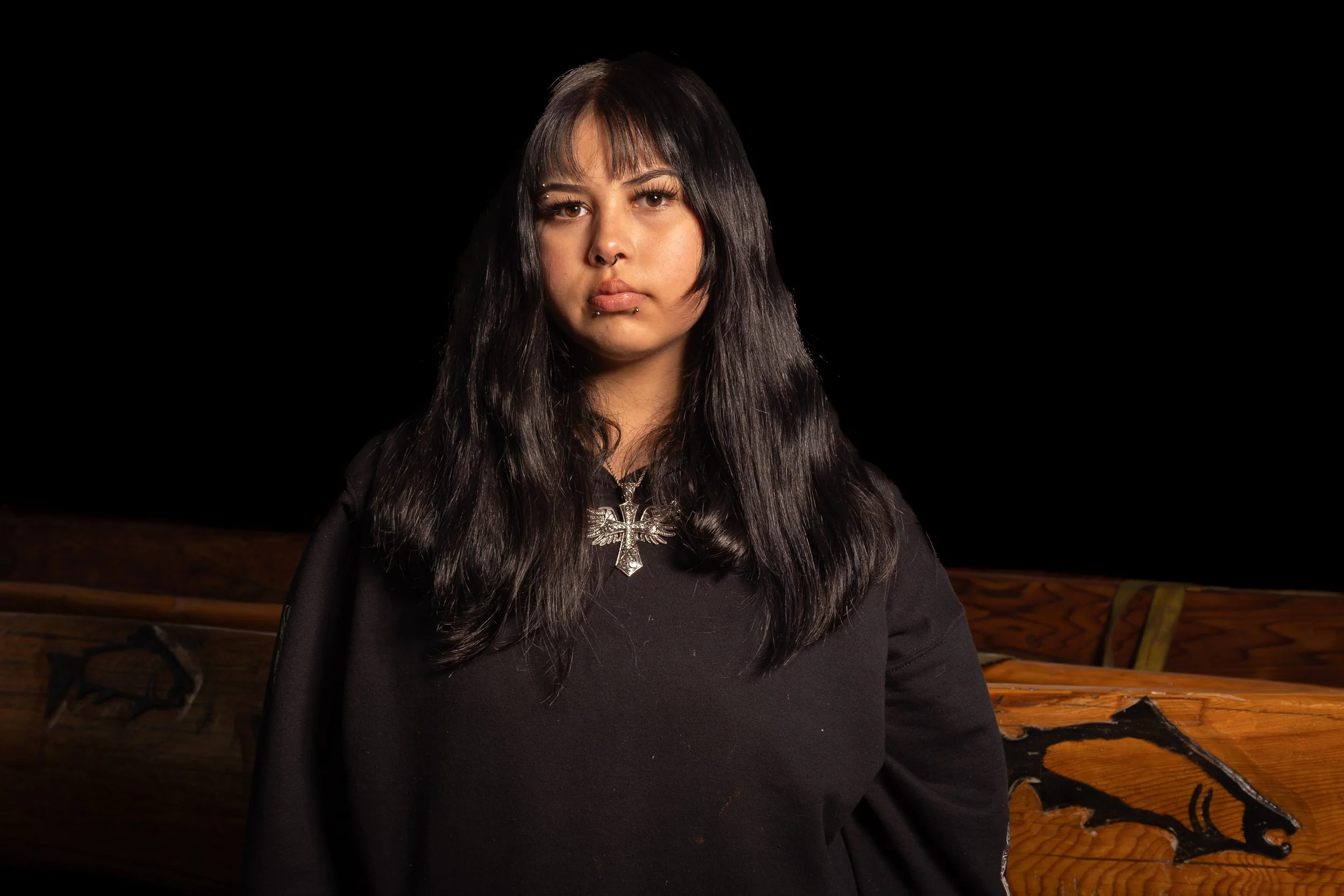 A woman with long black hair, piercings, and a cross necklace, wearing a black hoodie, standing indoors against a dark background.