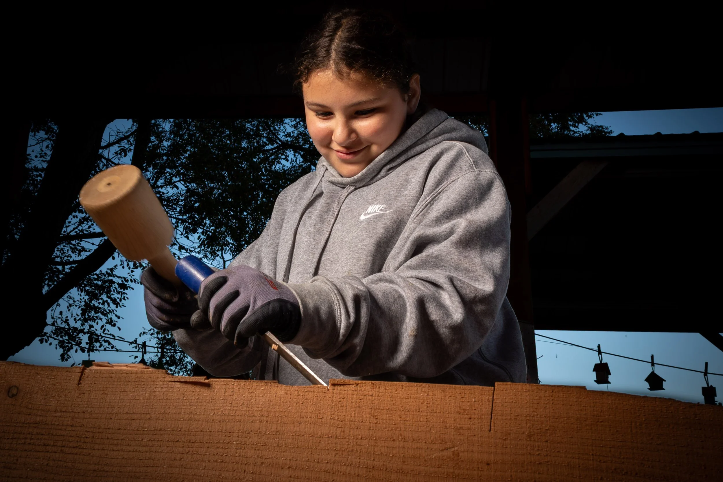A girl with brown hair in a ponytail wearing a gray Nike hoodie and gloves is using a hammer on a piece of wood outdoors during daytime. Trees and a building with a roof are visible in the background.