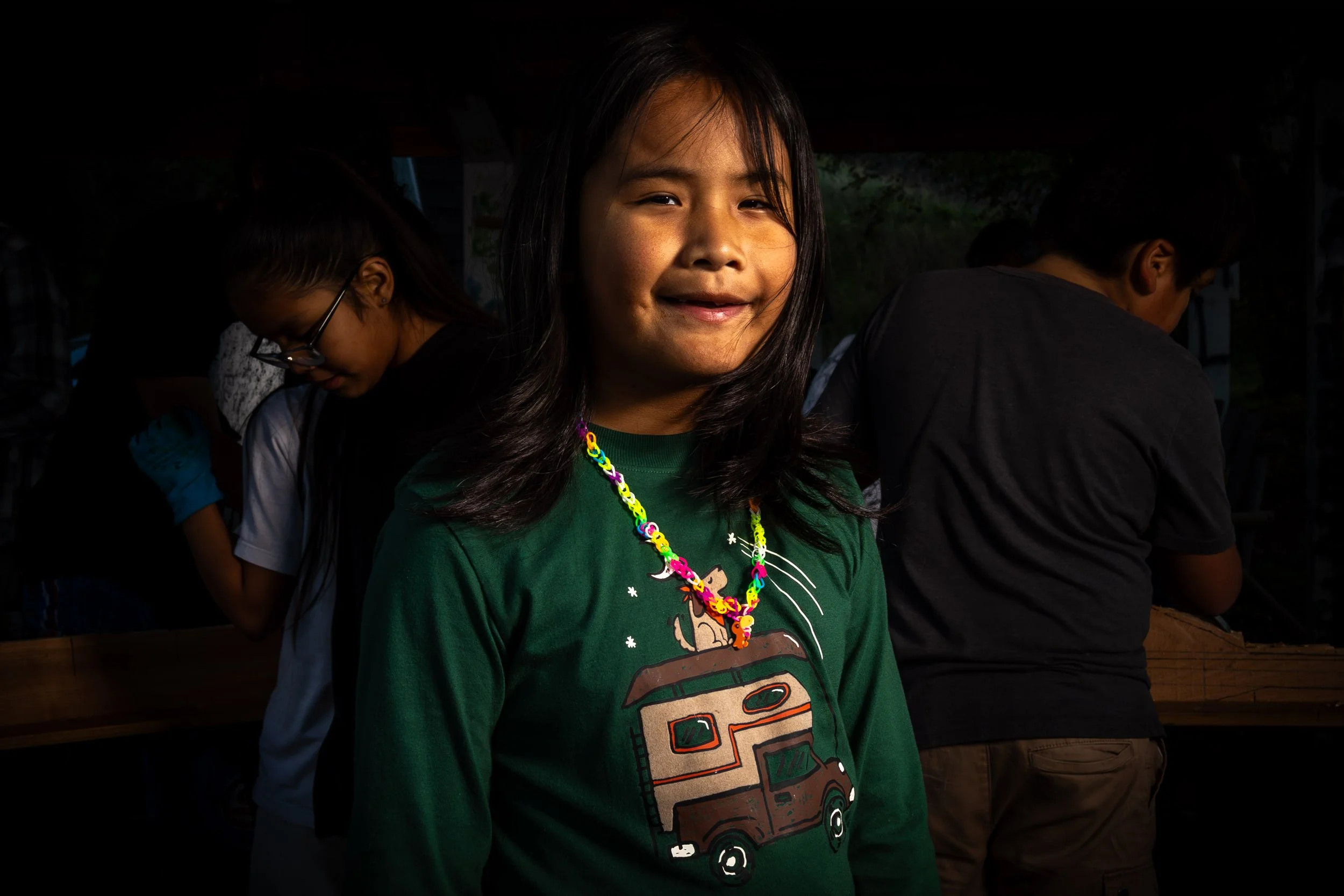 A young girl with dark hair smiling at the camera, wearing a green T-shirt and colorful beaded necklace, with other children in the background.