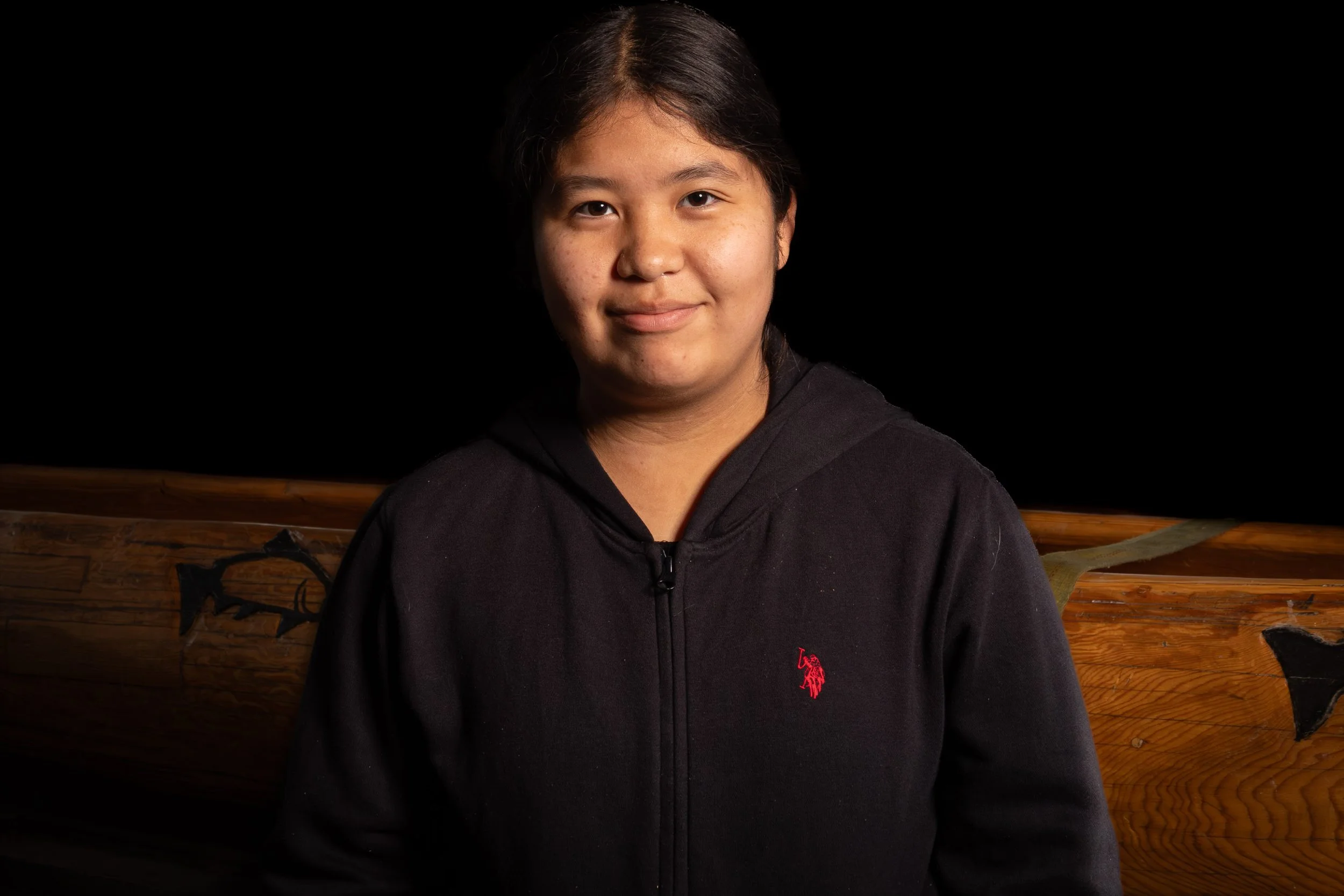 A young woman with dark hair, wearing a black jacket with red logo, standing in front of a wooden background.