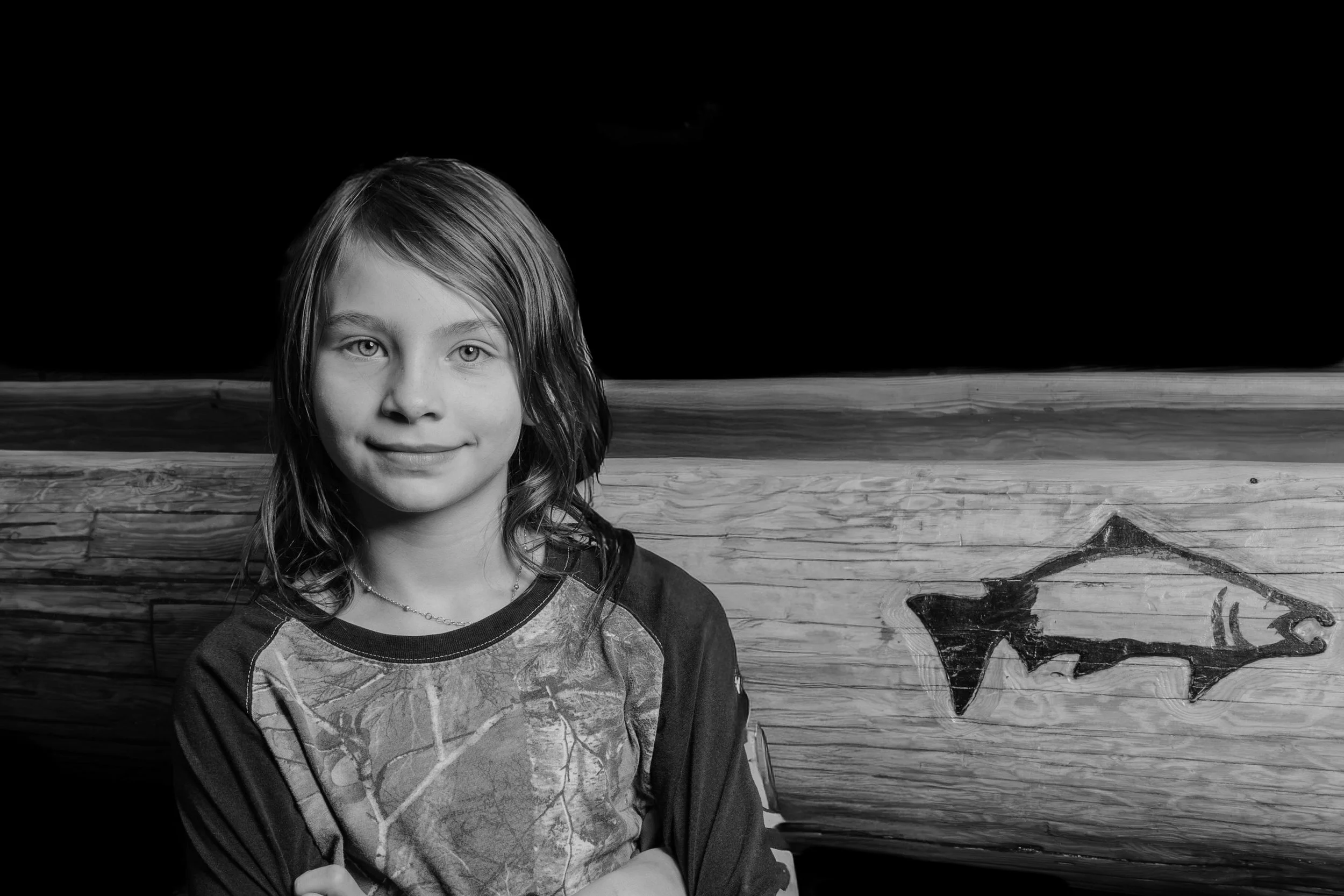 Black and white photo of a young girl with shoulder-length hair, wearing a patterned shirt and a necklace, standing in front of a wooden background with floral or animal illustrations.