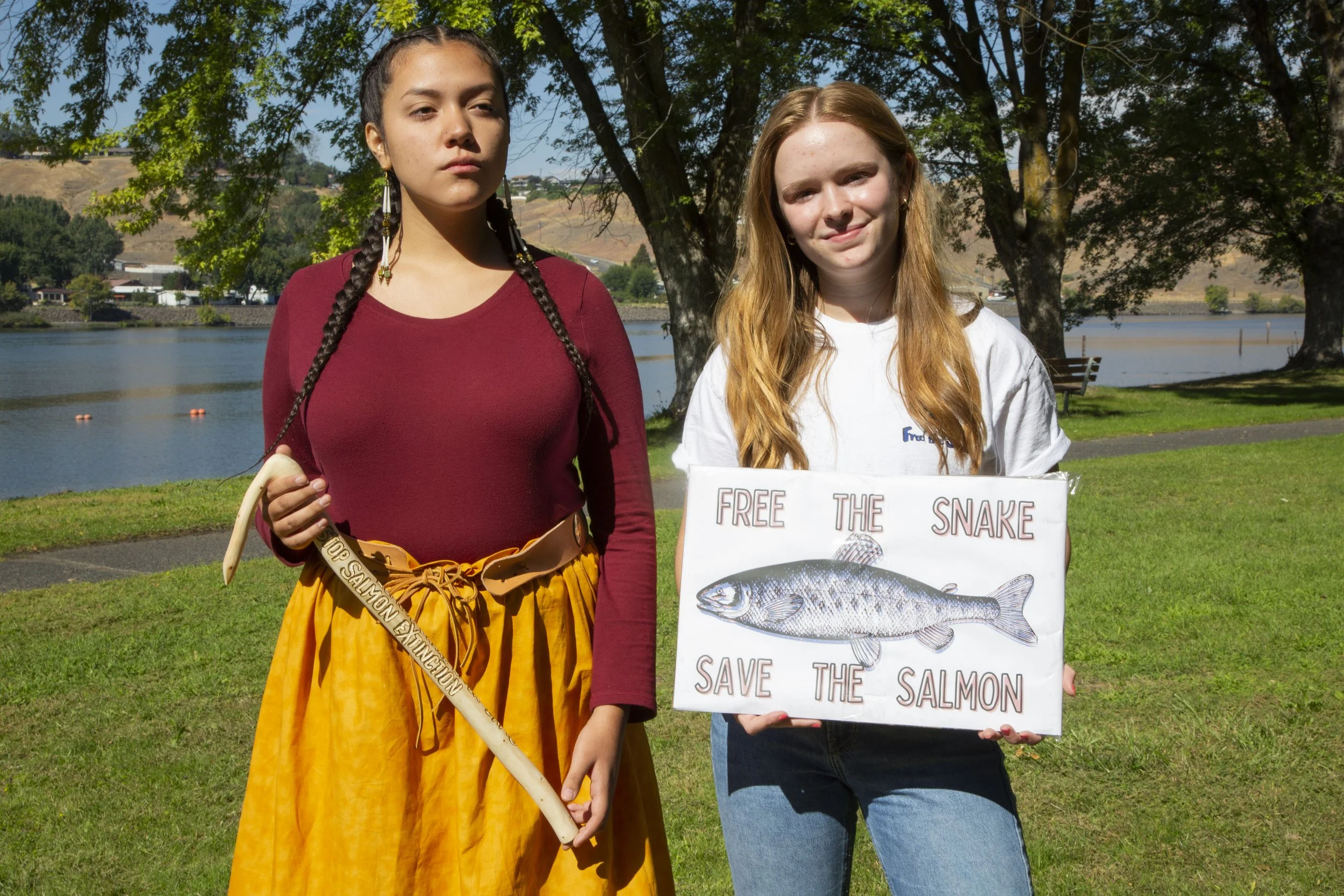 Two young women standing outdoors near a lake, one holding a sign that reads 'Free the Snake, Save the Salmon,' with trees and hills in the background.